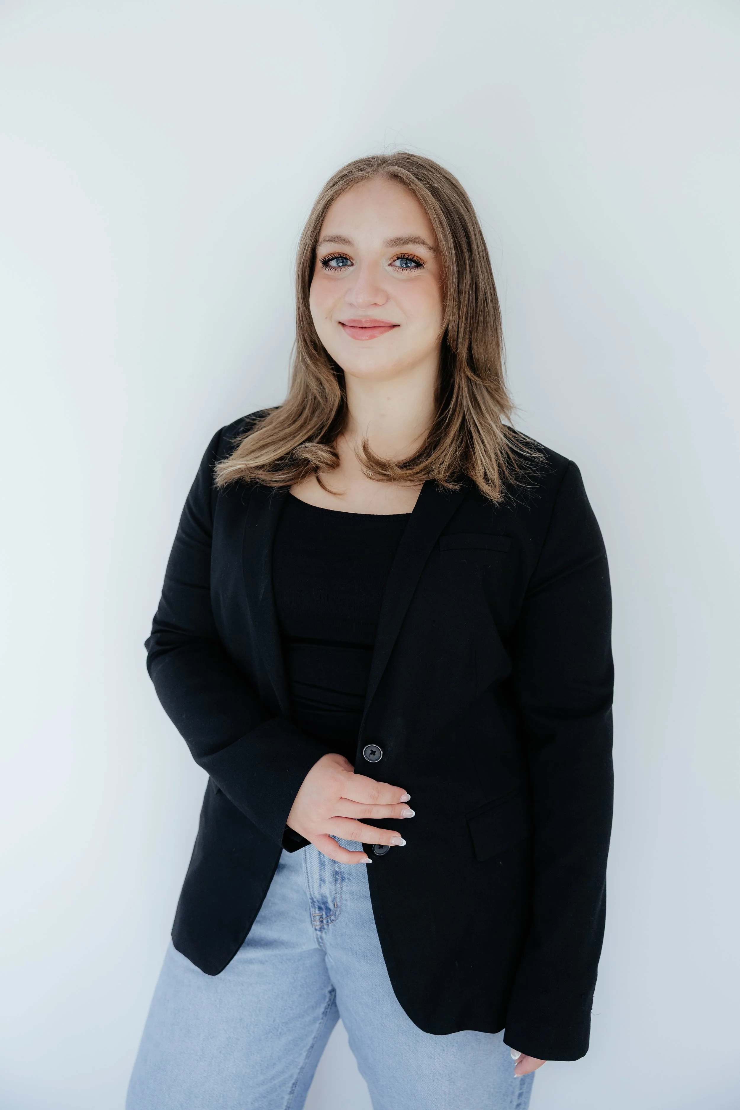 A young woman with shoulder-length light brown hair and blue eyes, smiling softly, wearing a black blazer over a black top, and light blue jeans, standing against a plain white background.