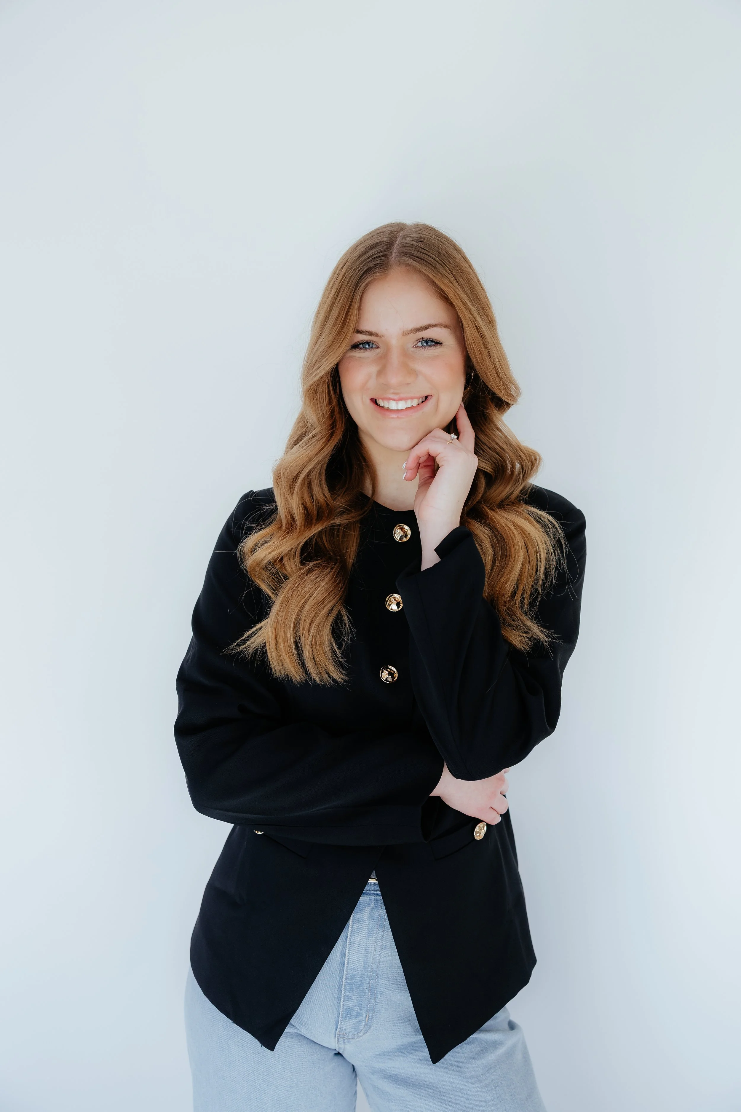A young woman with long wavy red hair, smiling and touching her chin with her right hand, wearing a black blazer with gold buttons and light blue jeans, standing against a plain white background.