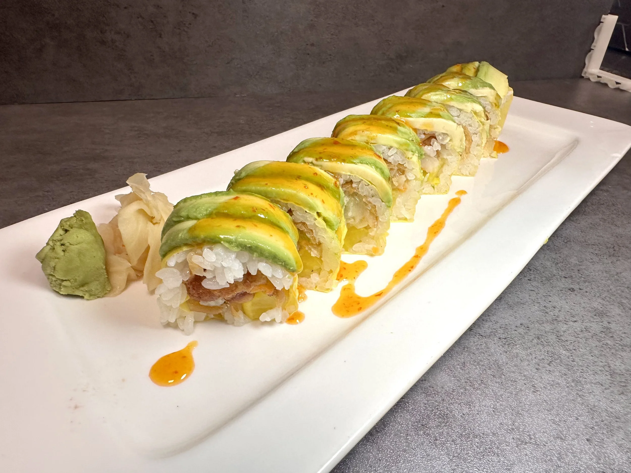 A plate of sushi rolls topped with sliced avocado and drizzled with orange sauce, accompanied by wasabi and pickled ginger.