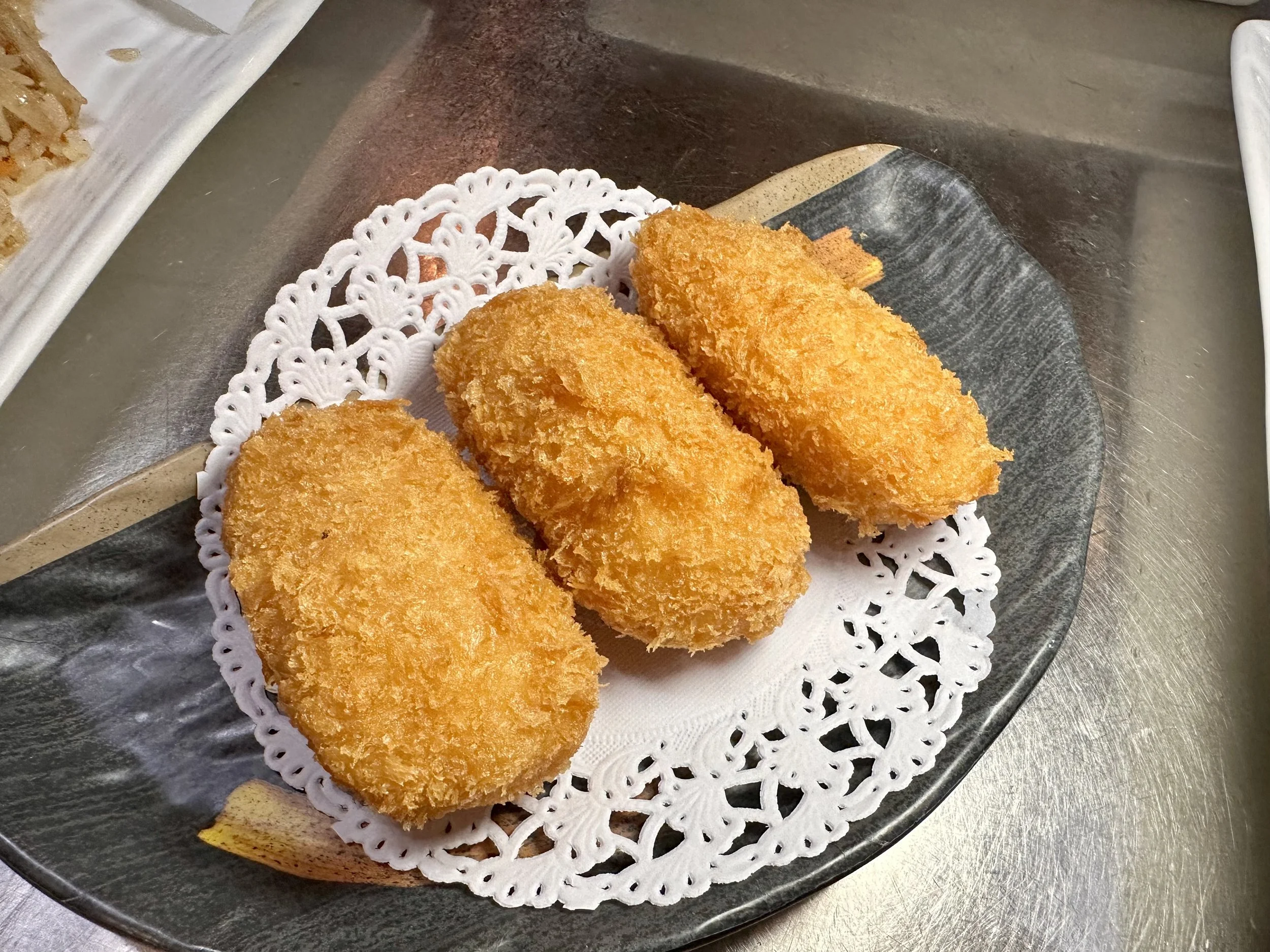 Four golden-brown breaded and fried oval-shaped croquettes served on a black ceramic plate with a decorative paper doily.