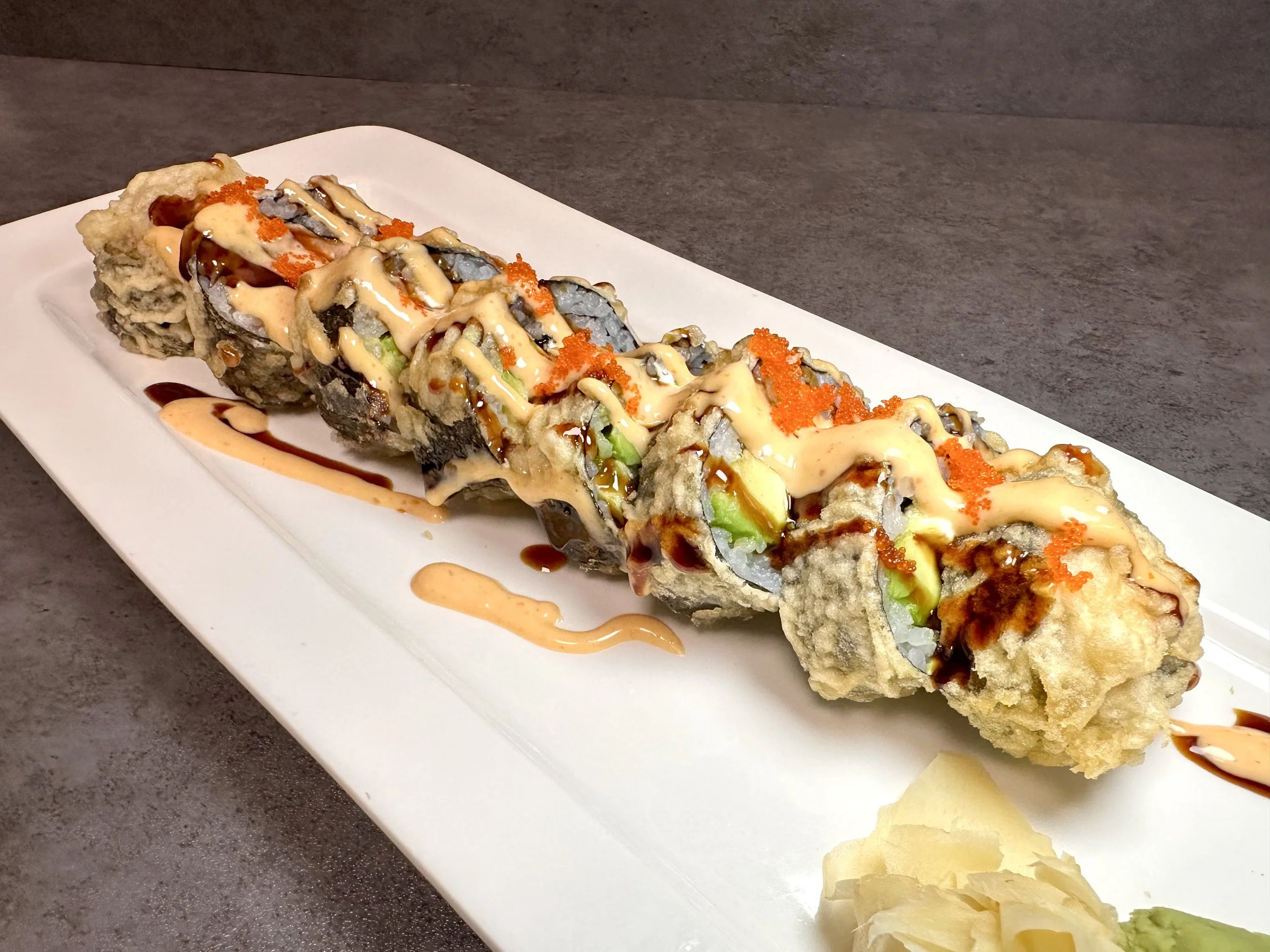 A plate of tempura sushi rolls topped with spicy mayo, eel sauce, and orange fish roe on a white rectangular plate.