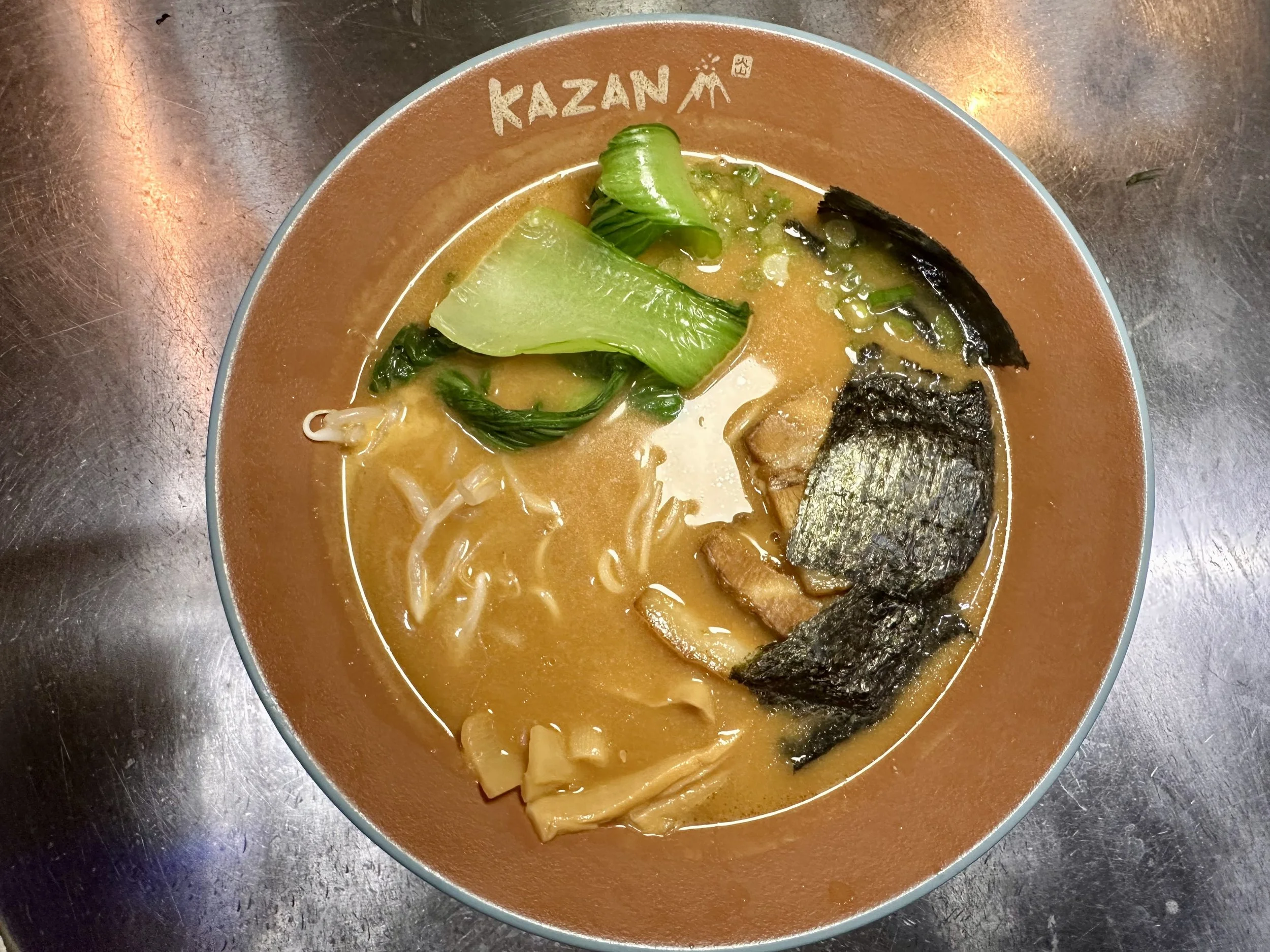 A bowl of ramen with seaweed, bok choy, and noodles in broth, on a metallic surface.