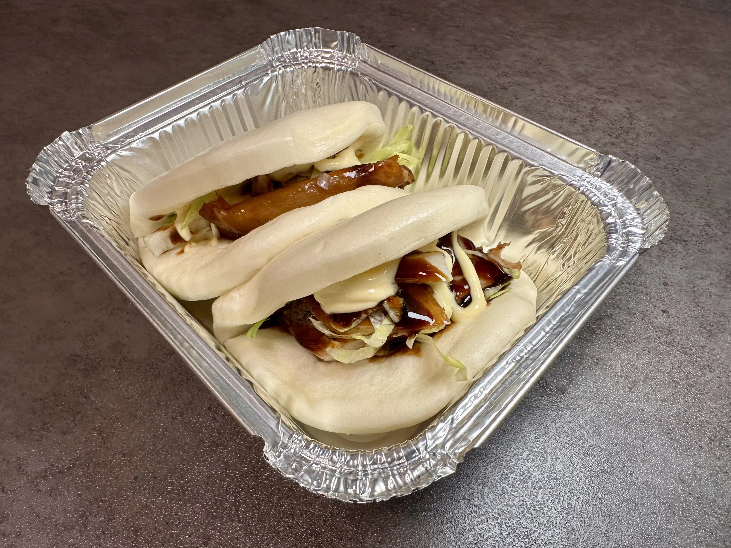Two steamed pork buns filled with pork, lettuce, and sauce in a foil container.