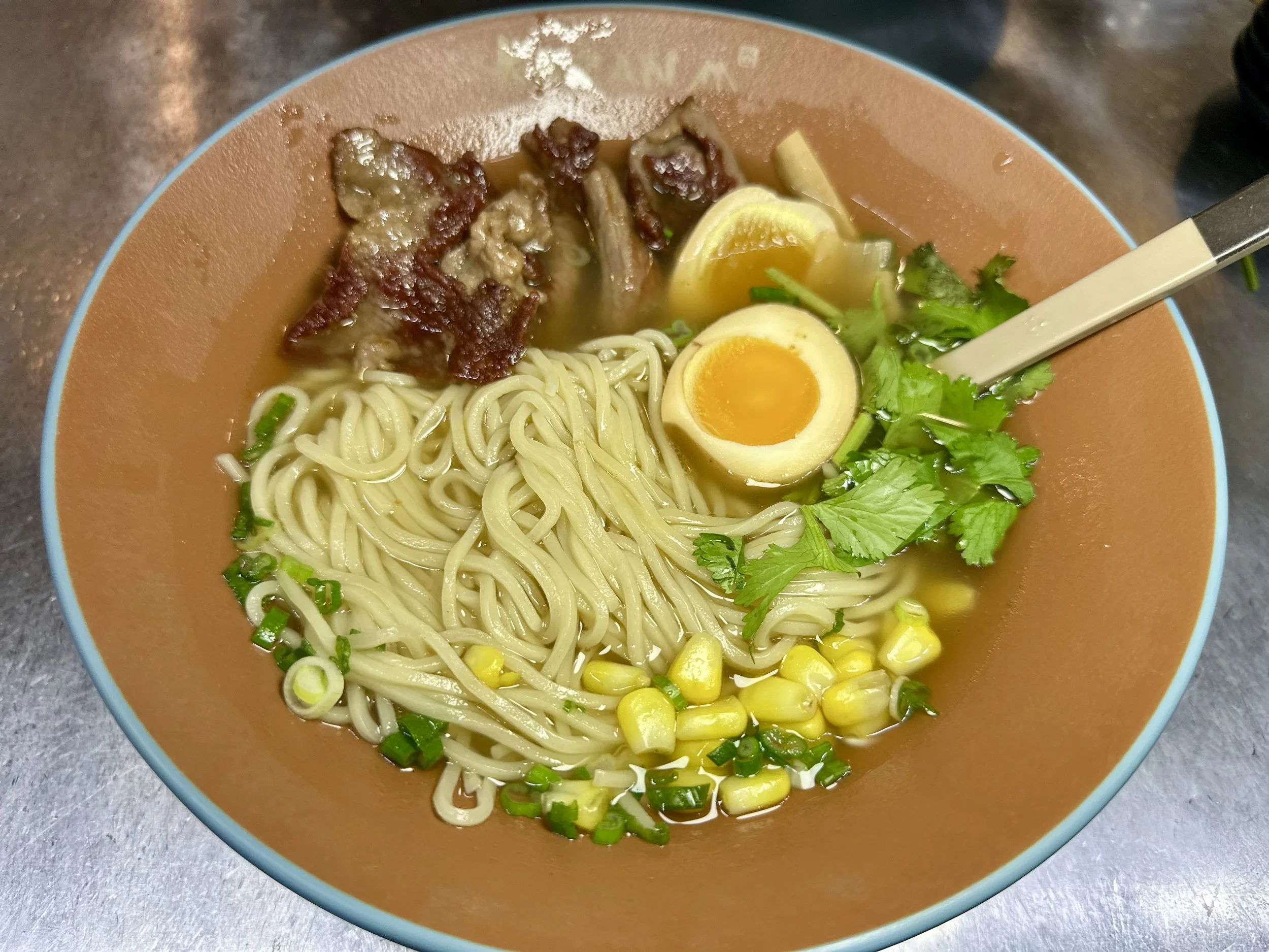 A bowl of ramen with noodles, sliced pork, a halved soft-boiled egg, corn, green onions, cilantro, lemon wedges, and broth.