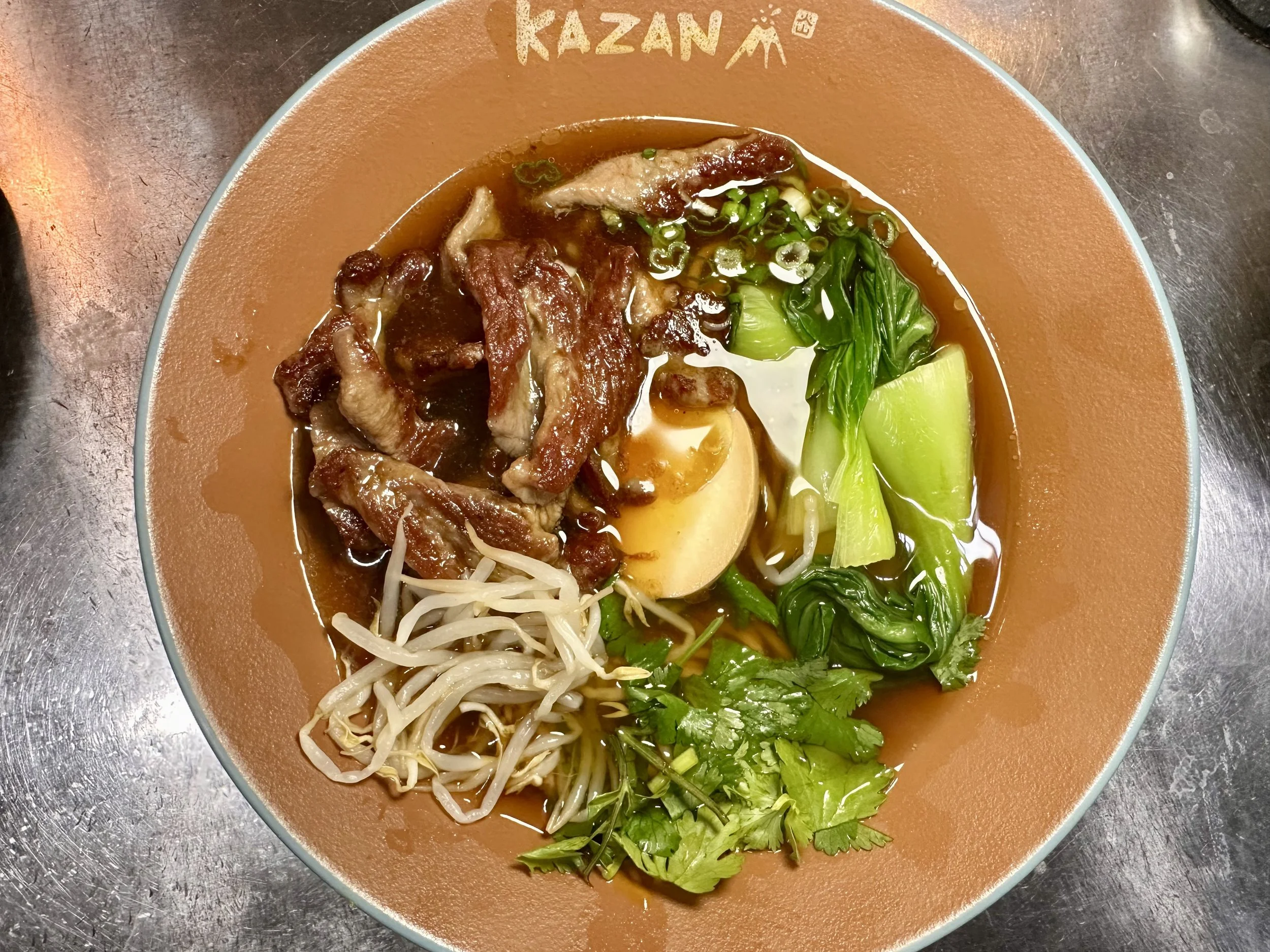 Bowl of ramen with slices of pork, a boiled egg, bean sprouts, chopped green onions, cilantro, and bok choy in broth on a brown plate labeled 'KAZAN'.