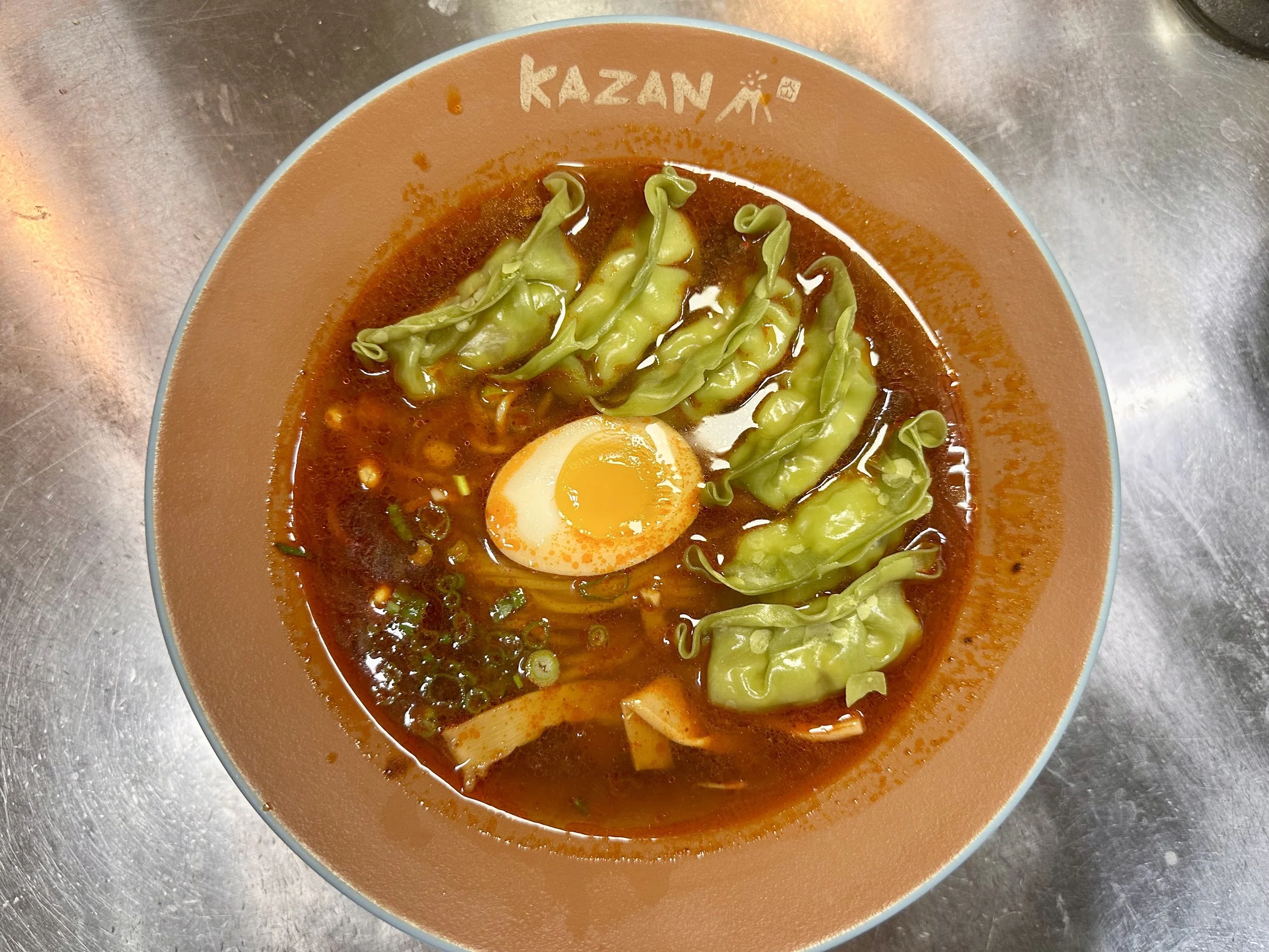 Bowl of ramen with sliced green vegetables, a soft-boiled egg, and broth on a metal surface.