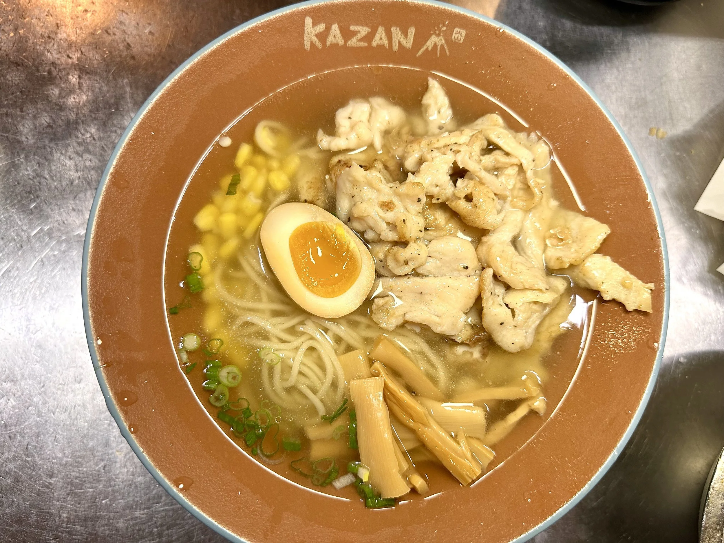 Bowl of ramen noodles with sliced green onions, a halved soft-boiled egg, sliced chicken, bamboo shoots, and corn, sitting on a metal surface