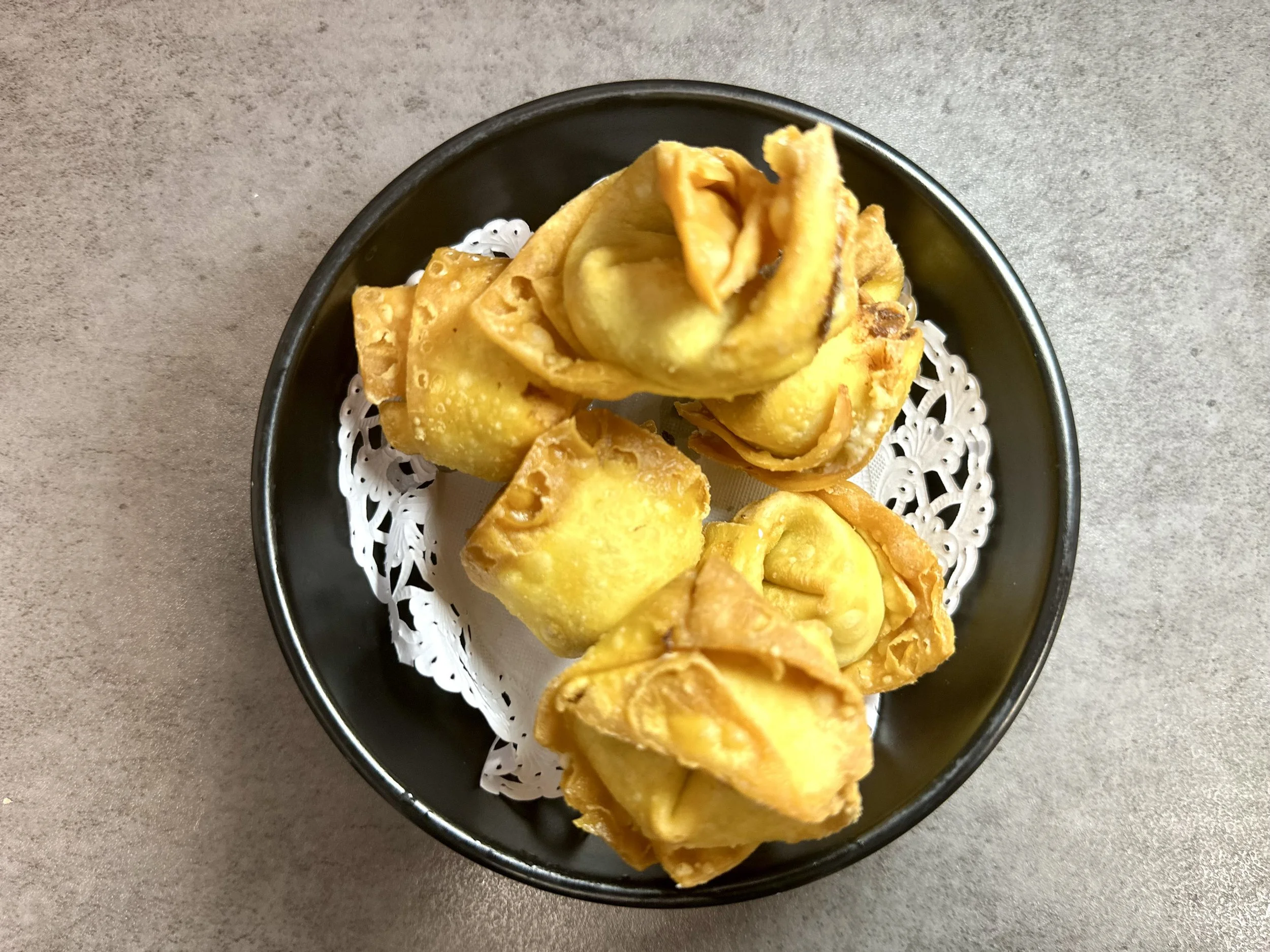 A black bowl with a paper doily containing six fried dumplings or egg rolls on a gray surface.