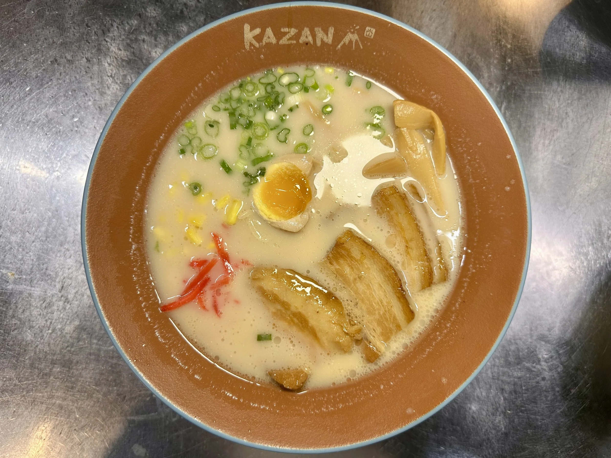 Bowl of ramen with broth, sliced pork, soft-boiled egg, chopped green onions, bamboo shoots, and red pickled ginger on a metal table.