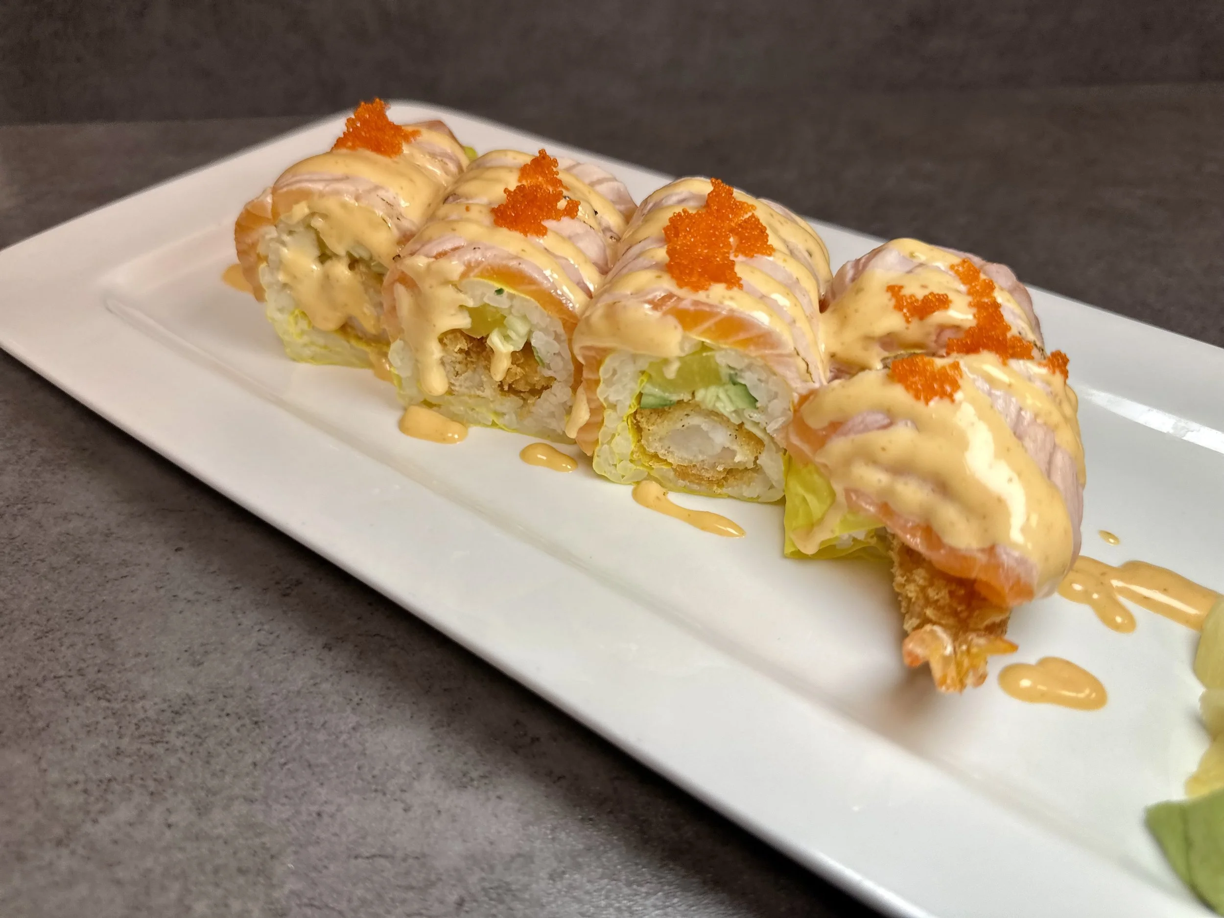 Sushi roll with salmon, lettuce, and a creamy sauce topped with orange fish roe on a white rectangular plate