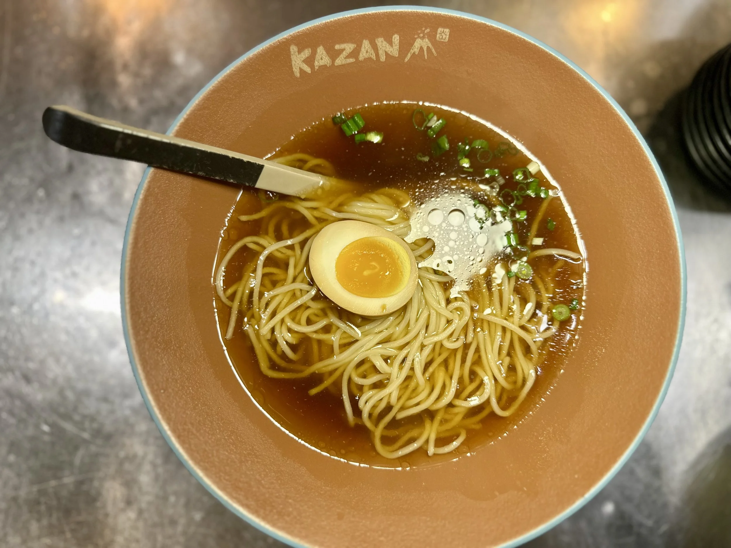 A bowl of ramen with noodles, a halved soft-boiled egg, chopped green onions, and broth, served in a bowl labeled 'Kazan'.