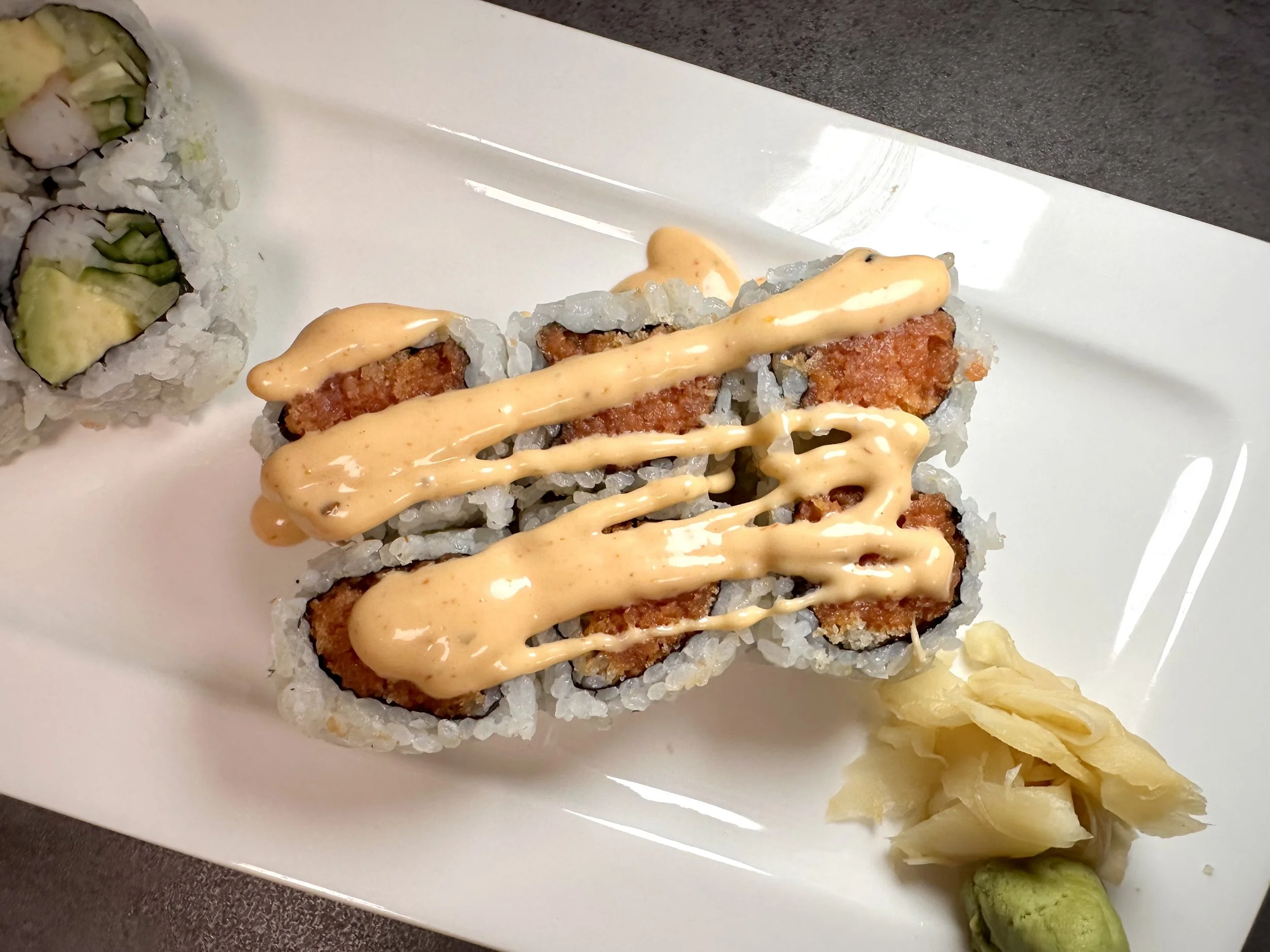 Sushi rolls with fried filling, topped with spicy mayo sauce, served with pickled ginger and wasabi on a white plate.