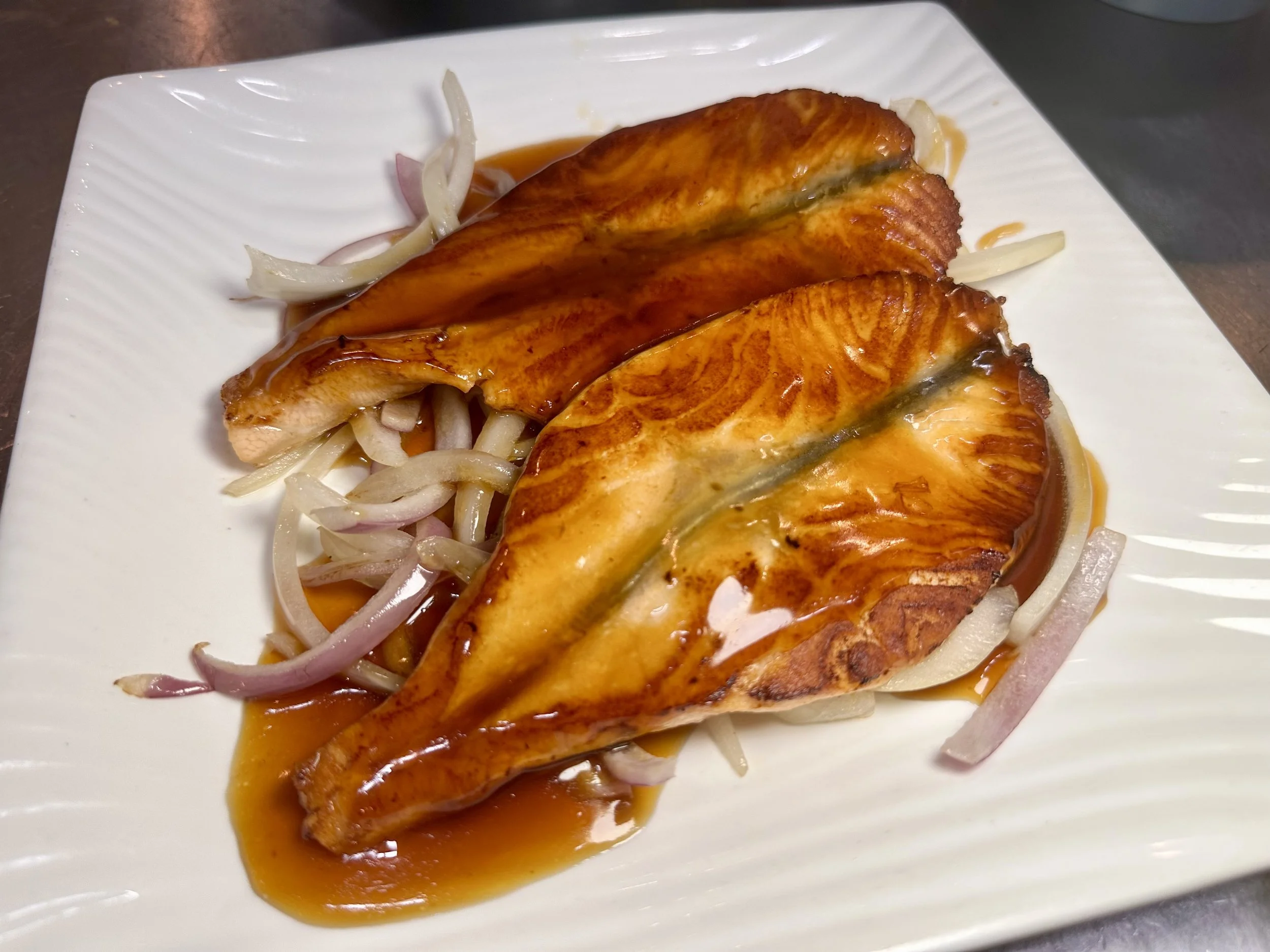 Three cooked salmon fillets with sauce on a white plate, garnished with sliced onions.
