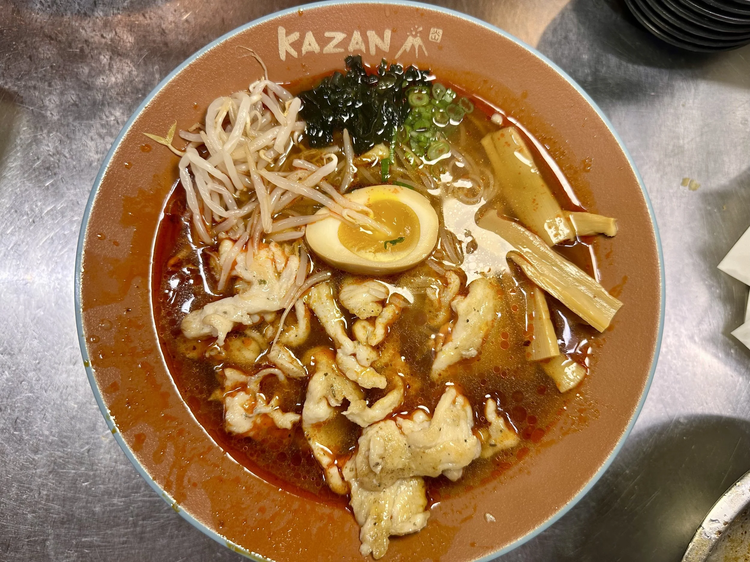 A bowl of spicy ramen with a soft boiled egg, bean sprouts, seaweed, green onions, bamboo shoots, and slices of meat, on a metal table.