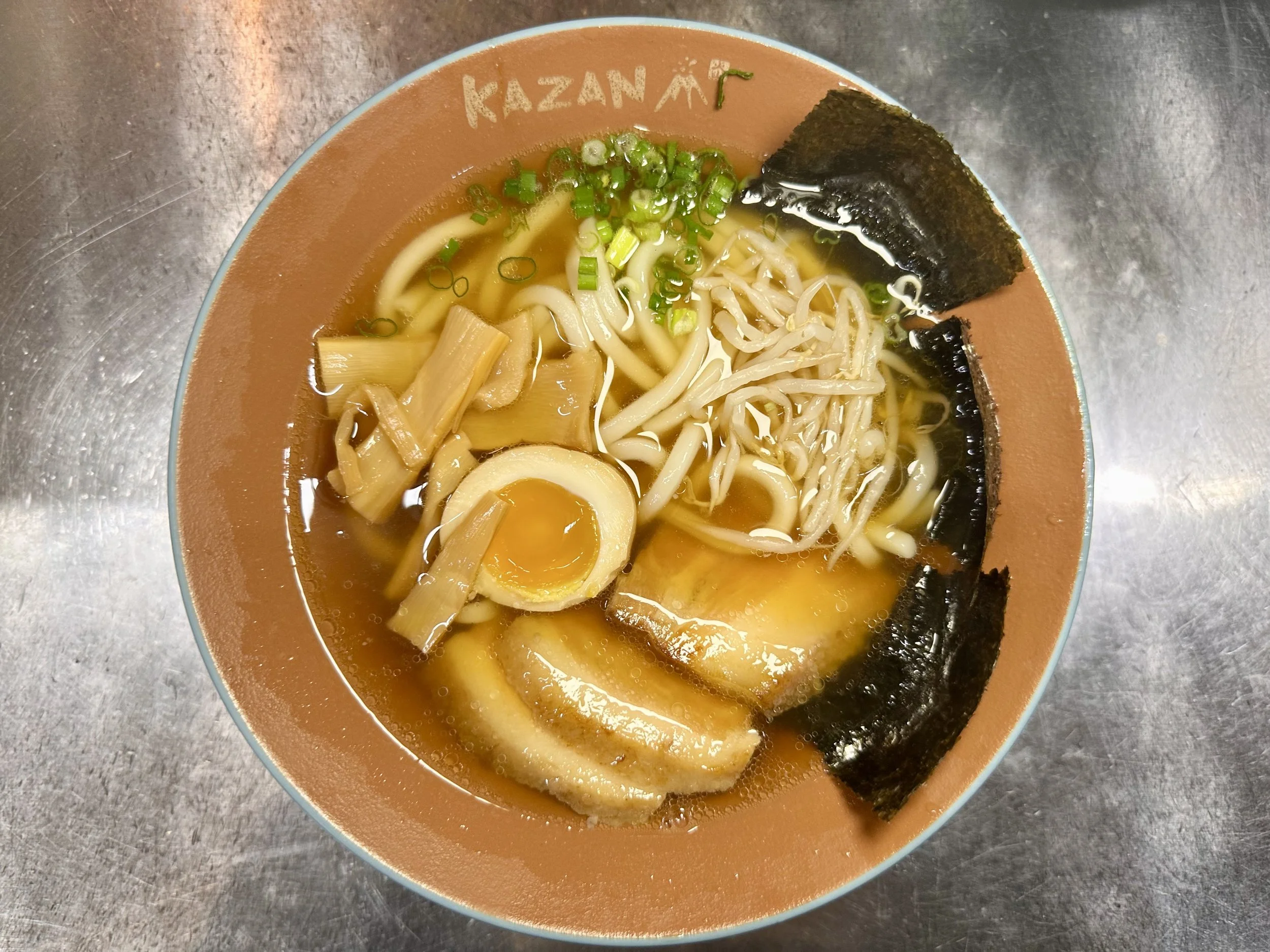 A bowl of ramen noodles with sliced pork, half a soft-boiled egg, bamboo shoots, chopped green onions, shredded chicken, and seaweed on top.