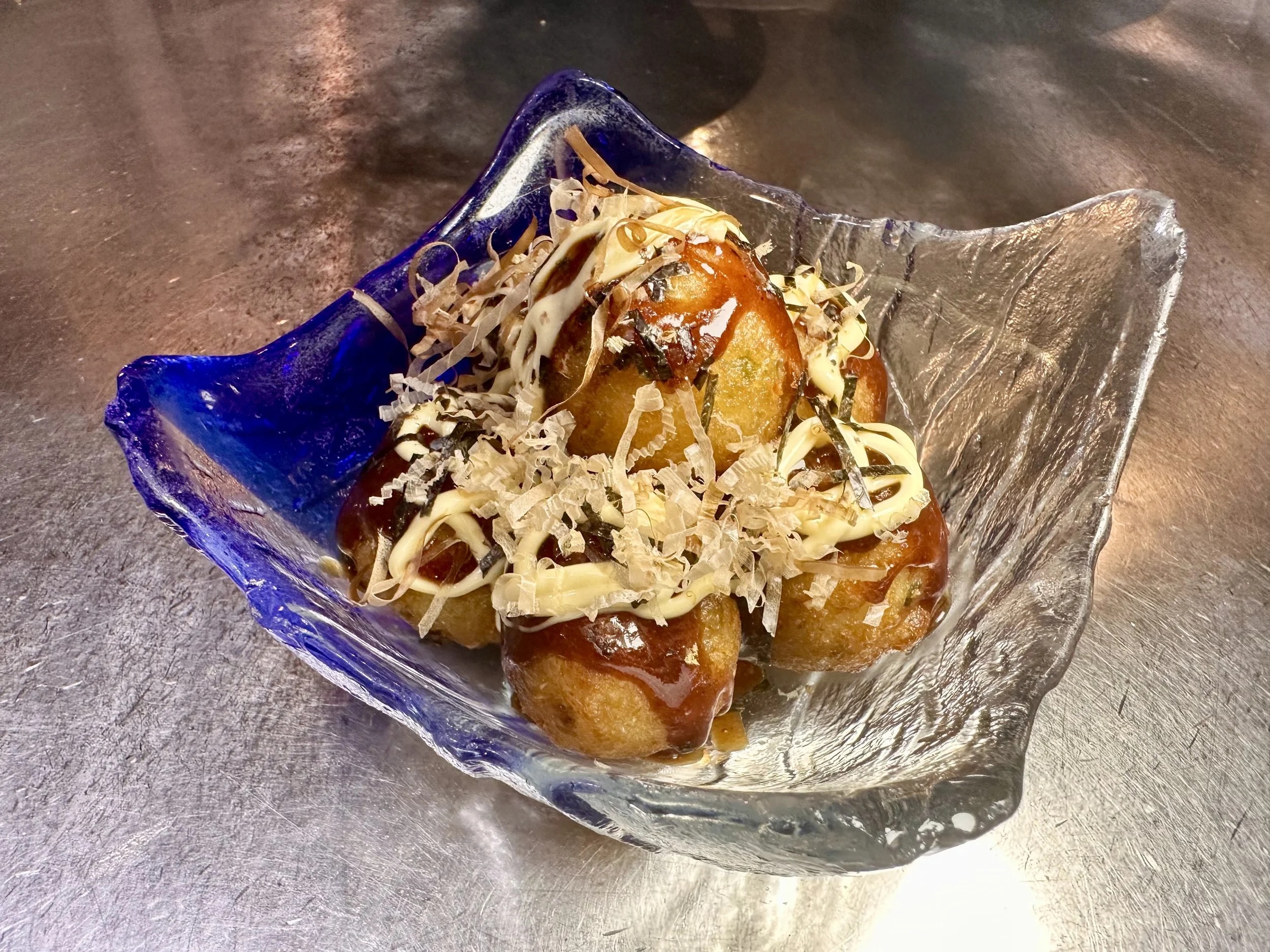 A glass dish with blue accents containing fried potato balls topped with shredded cheese, drizzled with white and dark sauces, garnished with herbs.