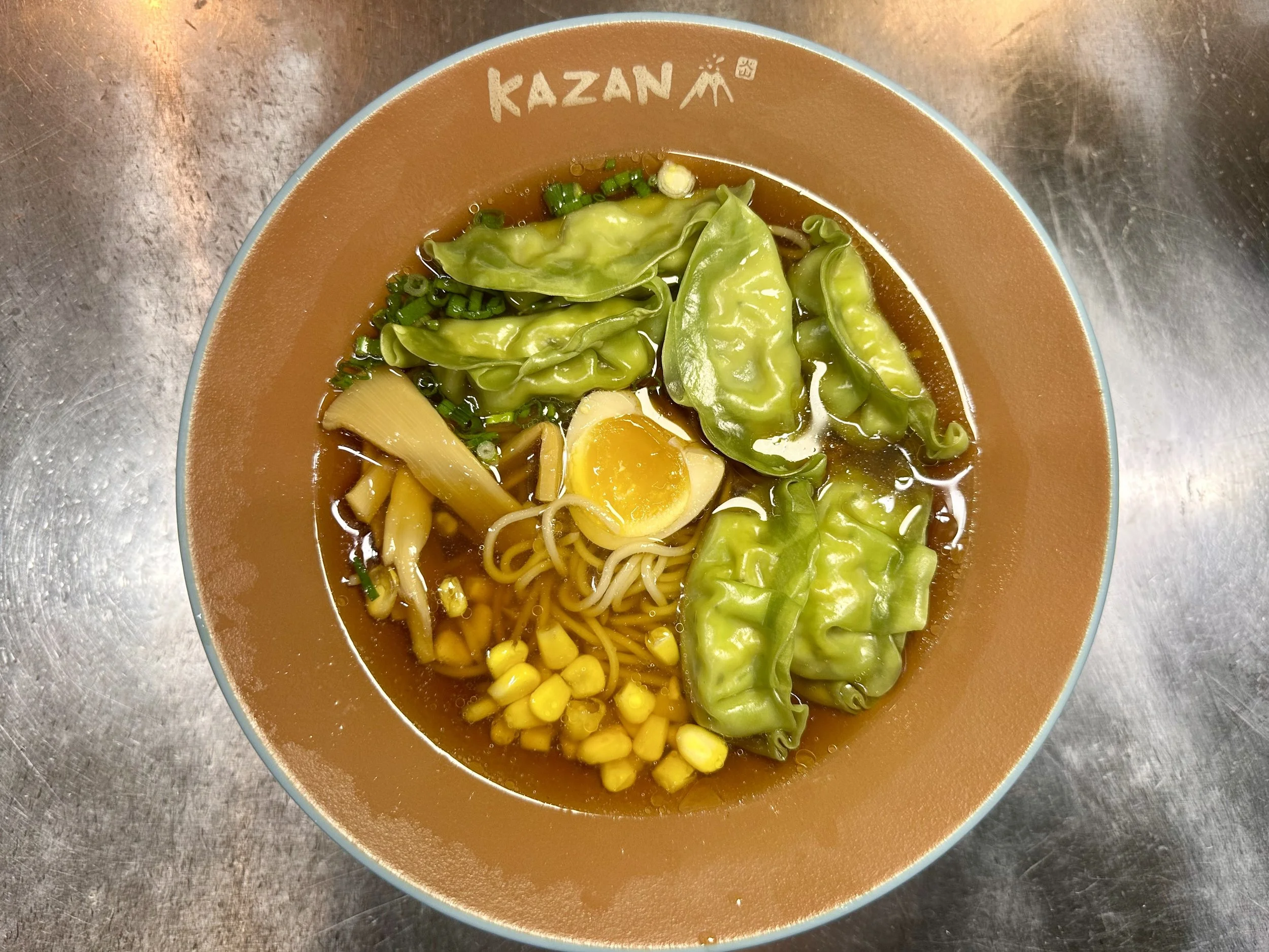 Bowl of ramen noodles topped with sliced green vegetables, corn, half an egg, and bamboo shoots, on a metallic surface.