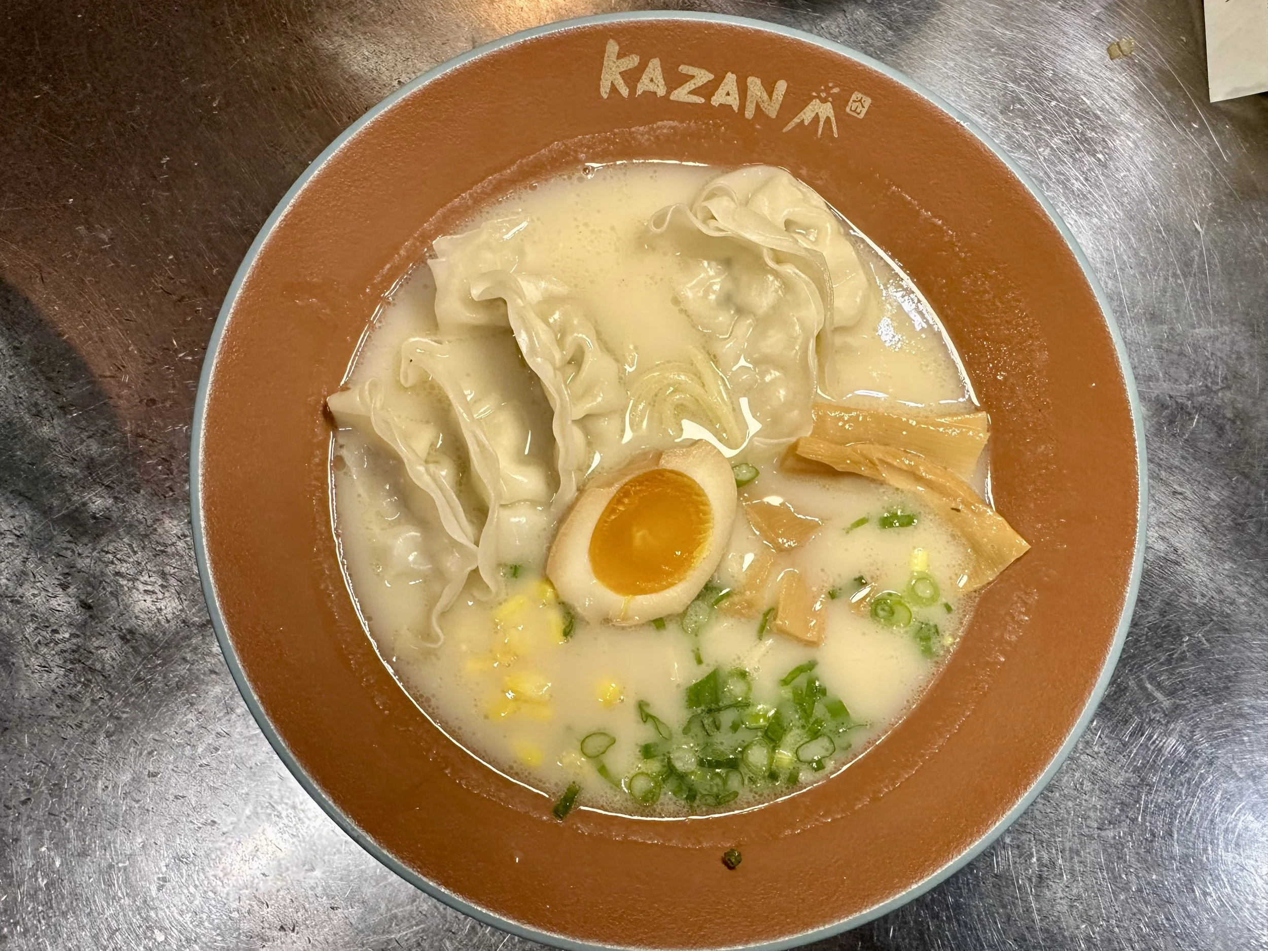 A bowl of ramen with egg, dumplings, green onions, and broth, on a metallic surface.