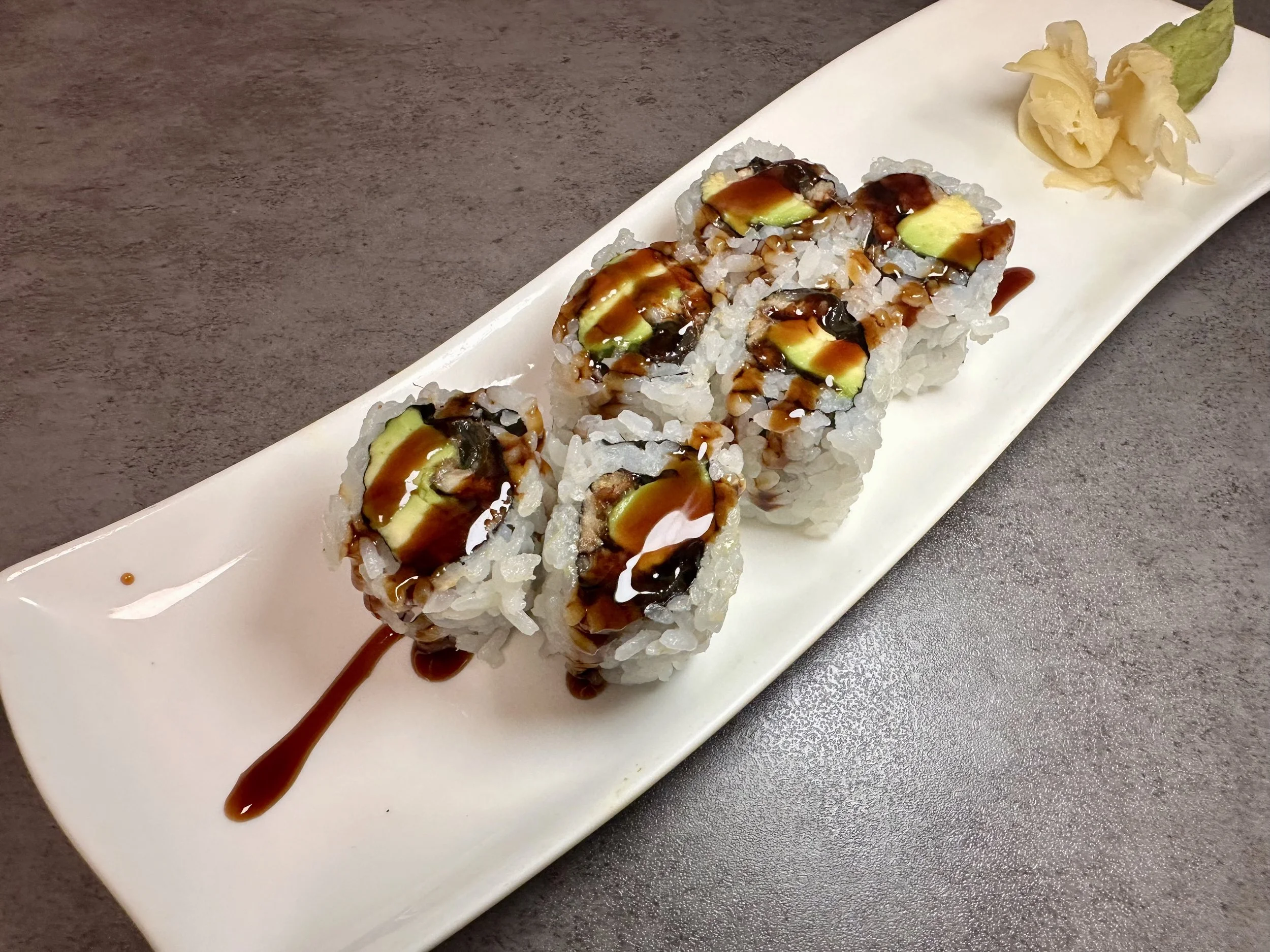 Sushi rolls topped with sauce on a white rectangular plate, garnished with pickled ginger and wasabi, placed on a gray surface.