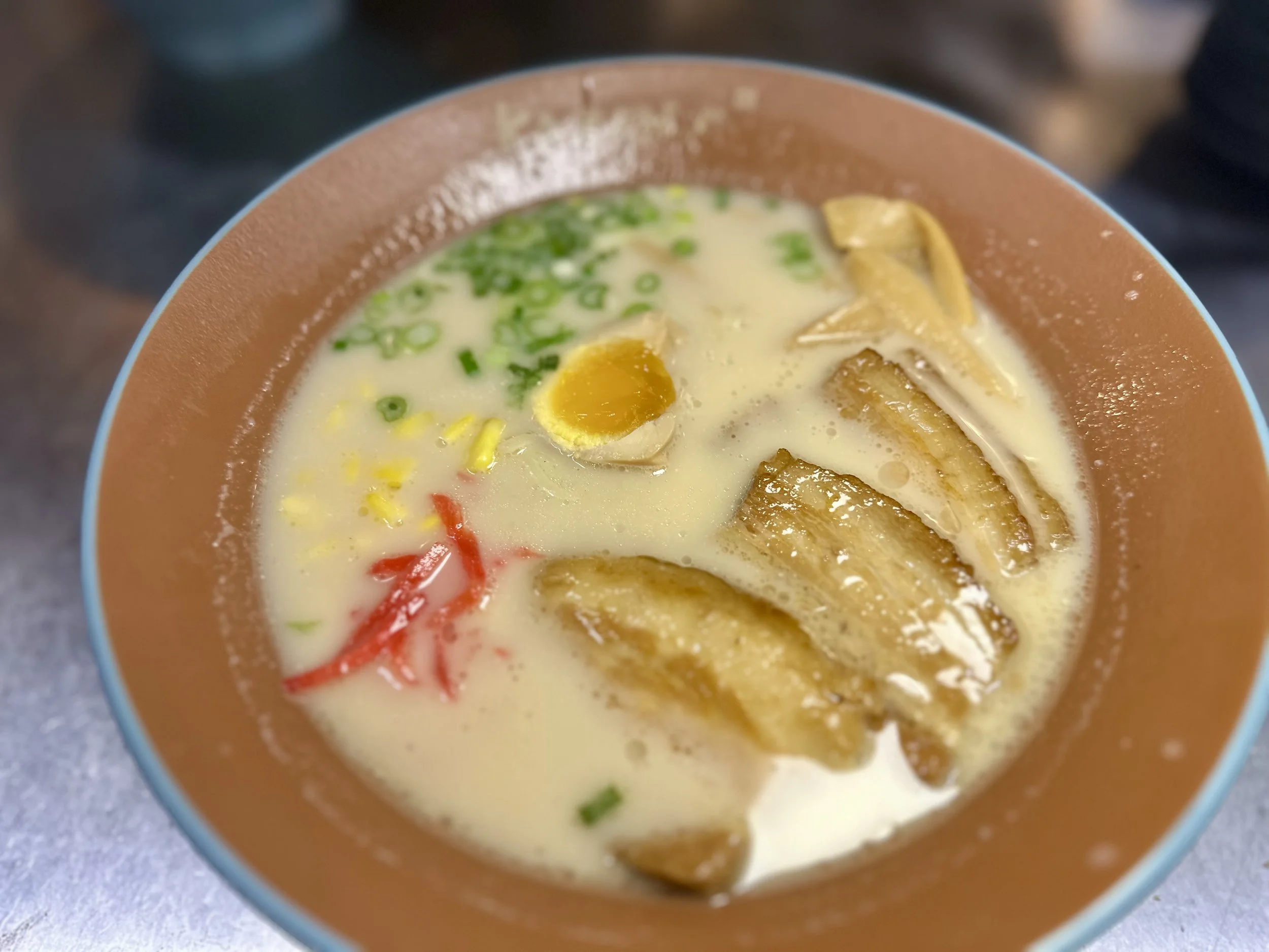 A bowl of ramen with creamy broth, slices of pork belly, chopped green onions, a half-boiled egg, and red and yellow garnishes.