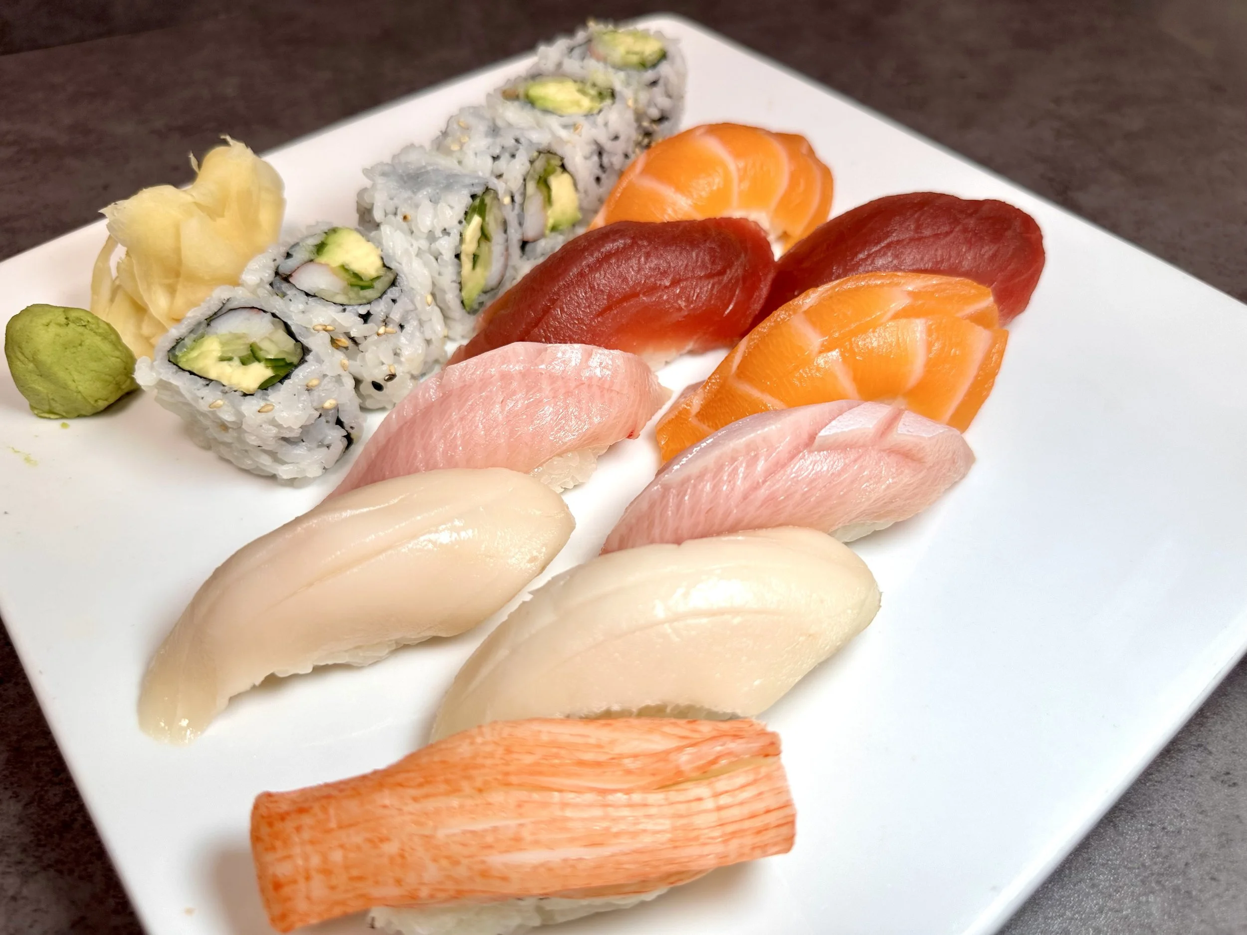 Assorted sushi pieces including tuna, salmon, white fish, and crab, with sushi rolls, wasabi, pickled ginger, and wasabi on a white plate.