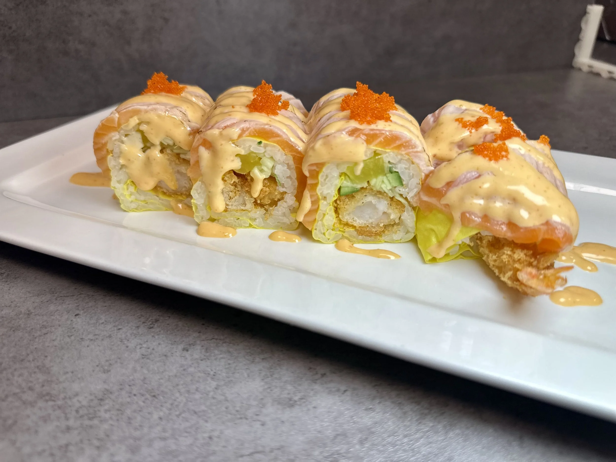 Sushi roll with rice, salmon, lettuce, and a creamy sauce, topped with orange fish roe on a white rectangular plate