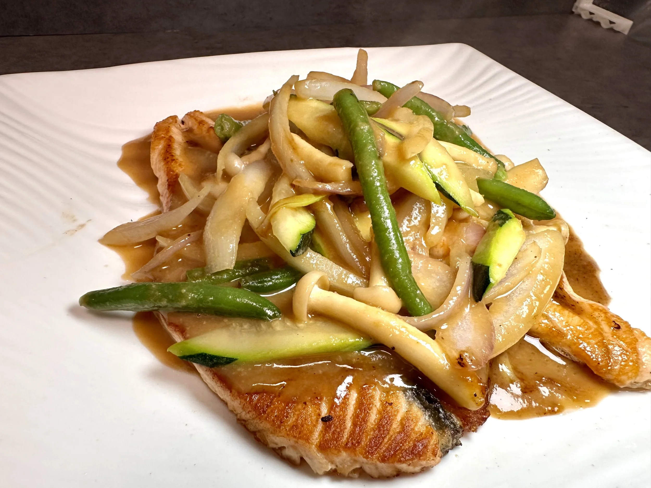 Grilled fish topped with a vegetable stir-fry including green beans, sliced zucchini, and onions, served with brown sauce on white plate.