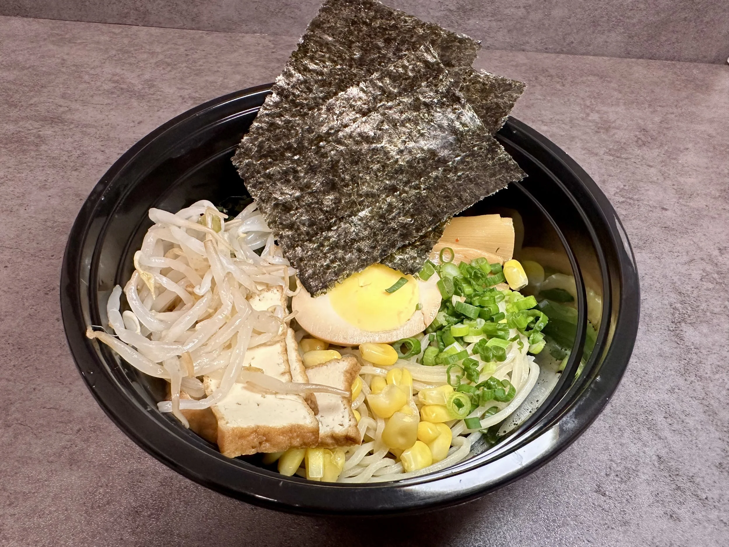 Bowl of ramen with seaweed, boiled egg, chopped green onions, corn, bean sprouts, and tofu on a gray countertop.