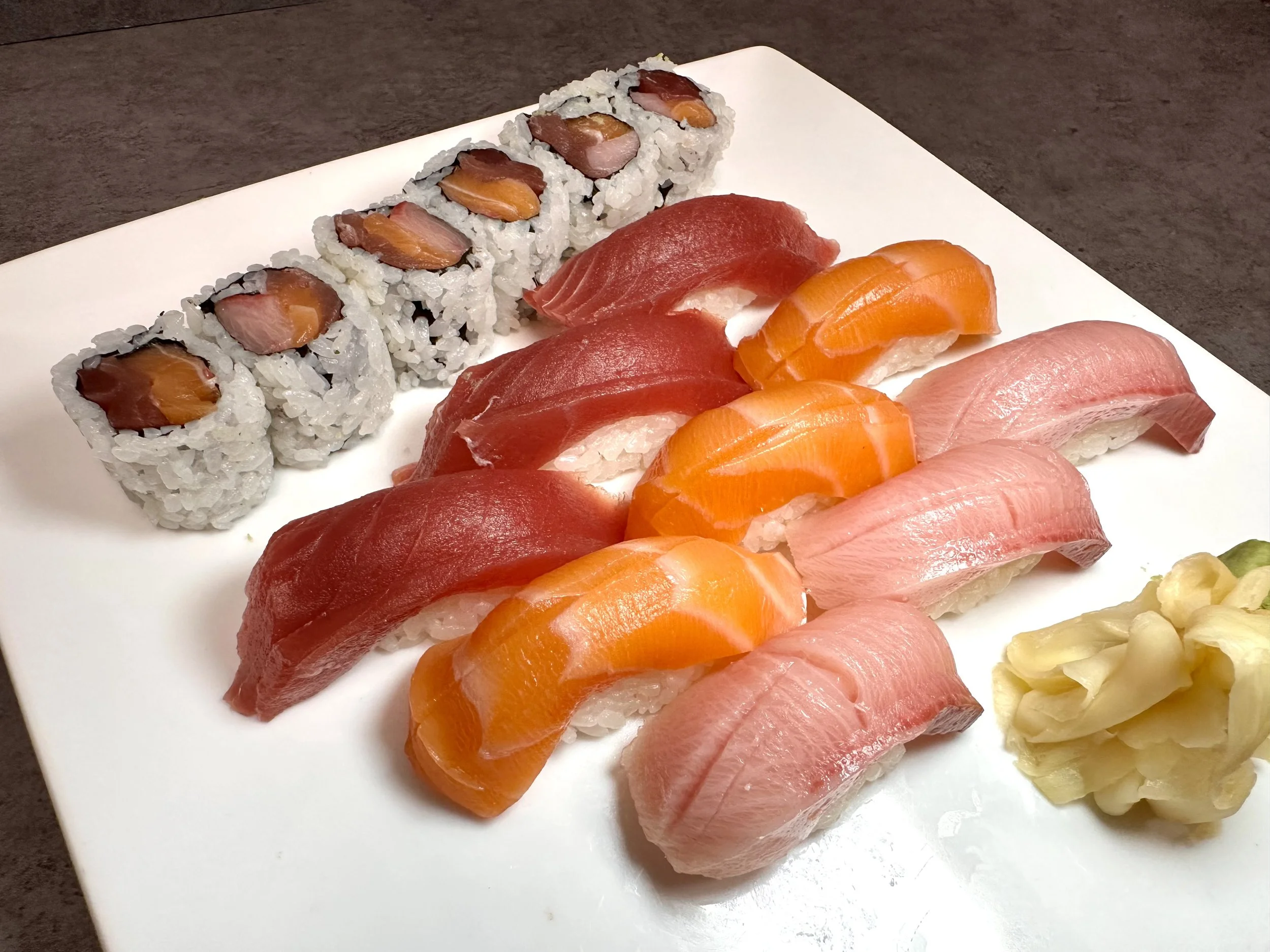 Assorted sushi including salmon and tuna nigiri, salmon and tuna sashimi, and a roll with fish inside, served with pickled ginger on a white plate.