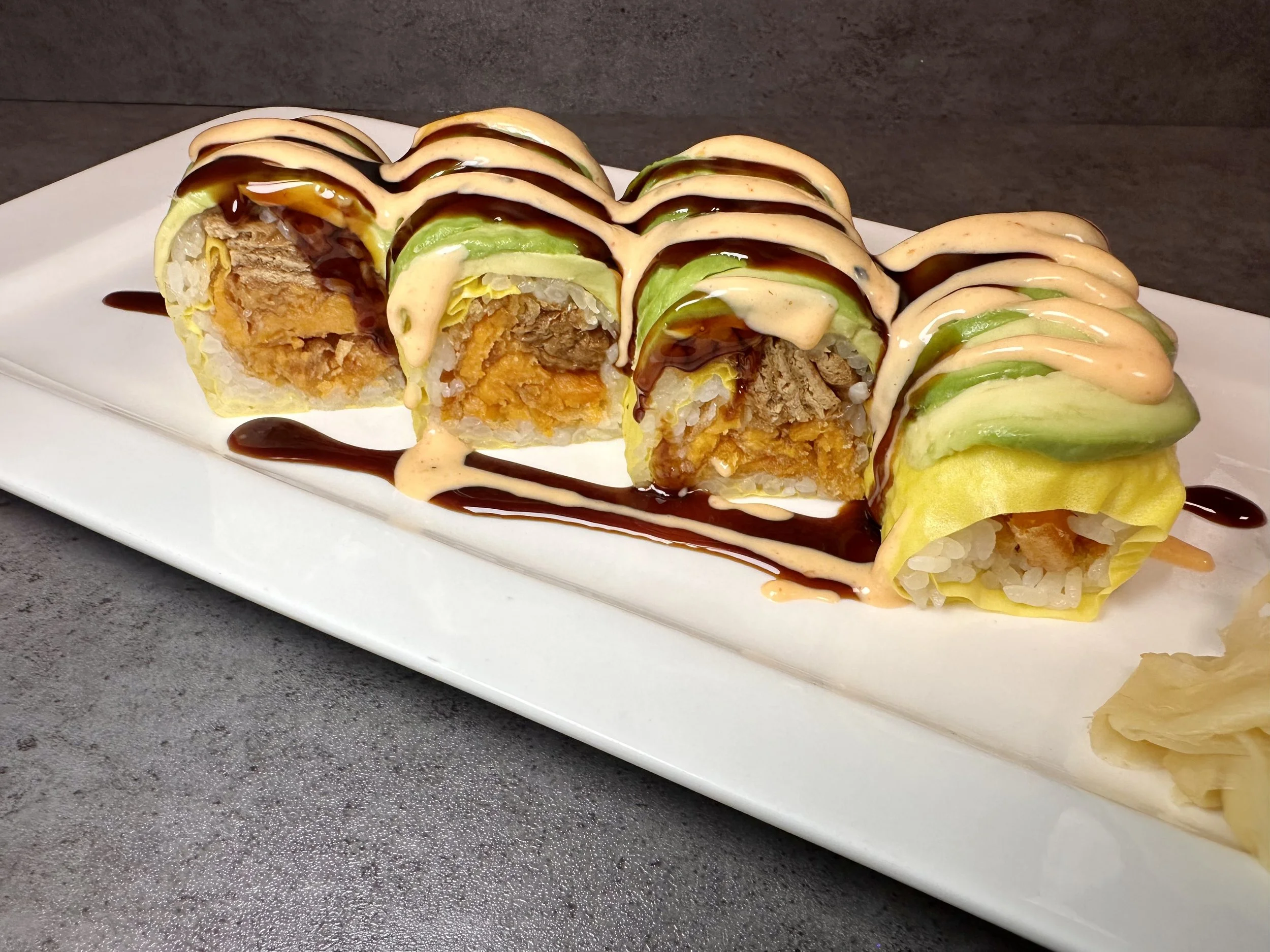 Close-up of a sushi roll on a white rectangular plate, topped with drizzles of dark and light sauces, with some ginger on the side, placed on a dark gray surface.