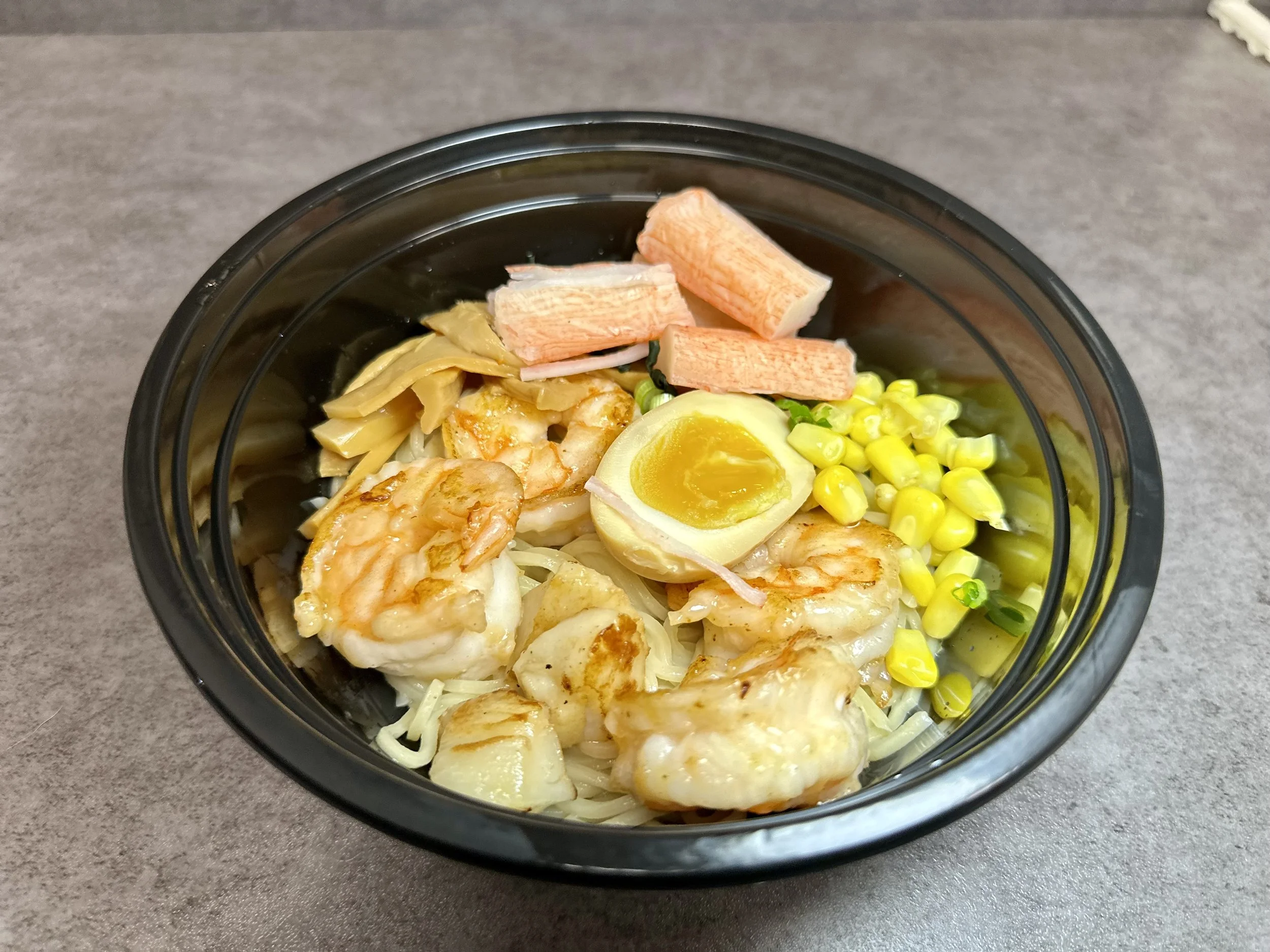 A black bowl containing a shrimp salad with boiled egg half, canned corn, shredded chicken, shredded cheese, crab sticks, and chopped green onions on a gray surface.