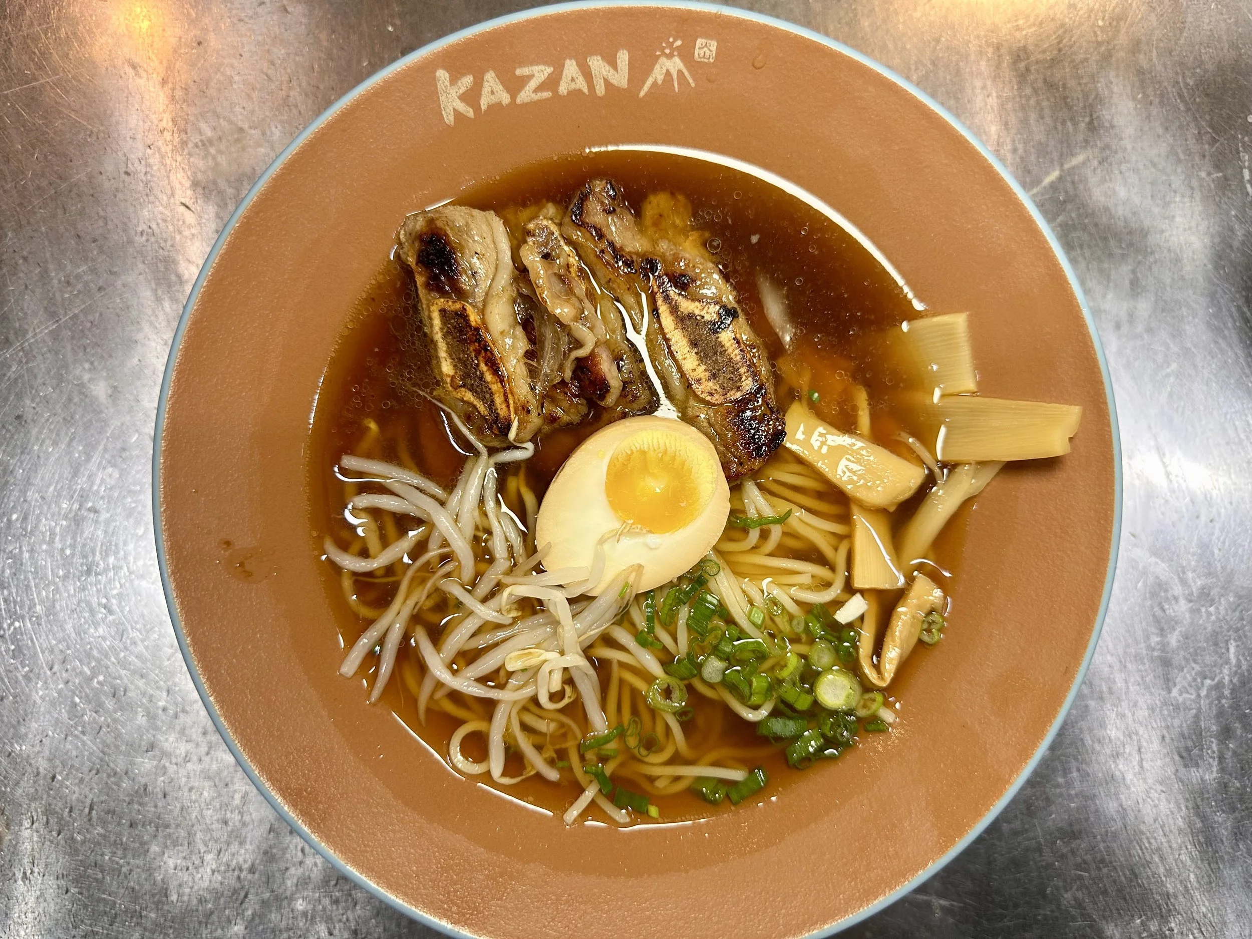 Bowl of ramen with grilled pork, half boiled egg, bean sprouts, chopped green onions, bamboo shoots, and noodles in broth, on a metallic surface.