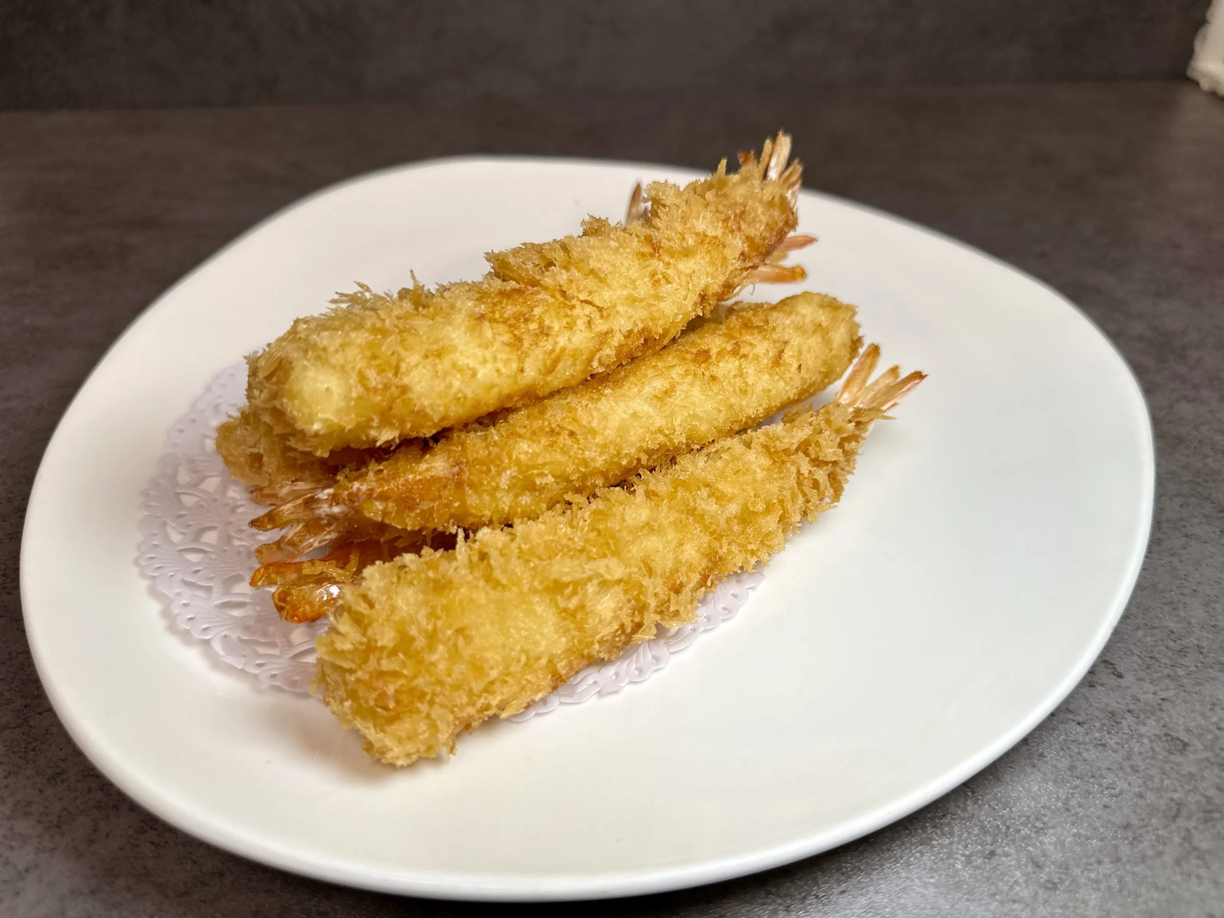 Three pieces of golden-brown breaded fried shrimp on a white plate with a paper doily, set against a dark surface.