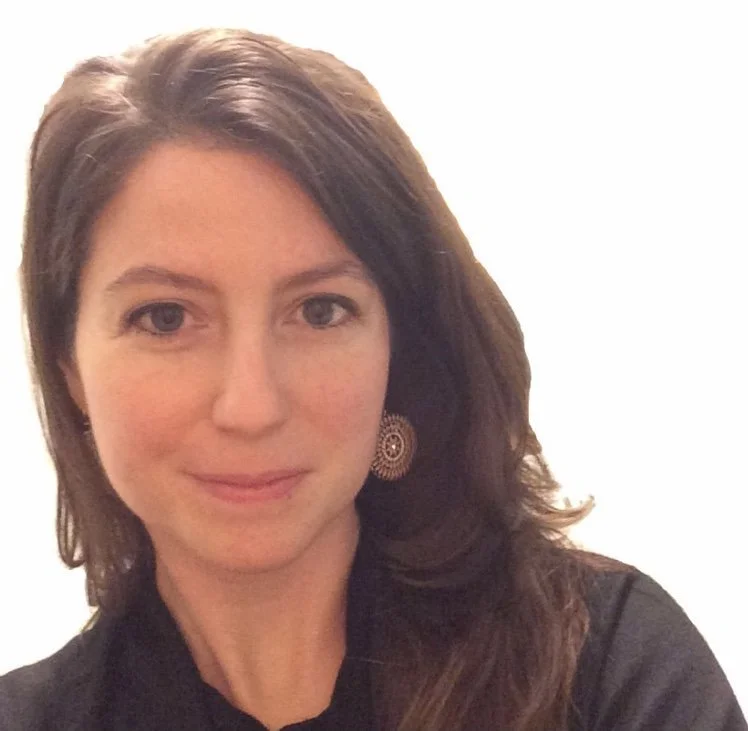 Close-up of a woman with shoulder-length brown hair, wearing a black top and decorative earrings, against a plain light background.