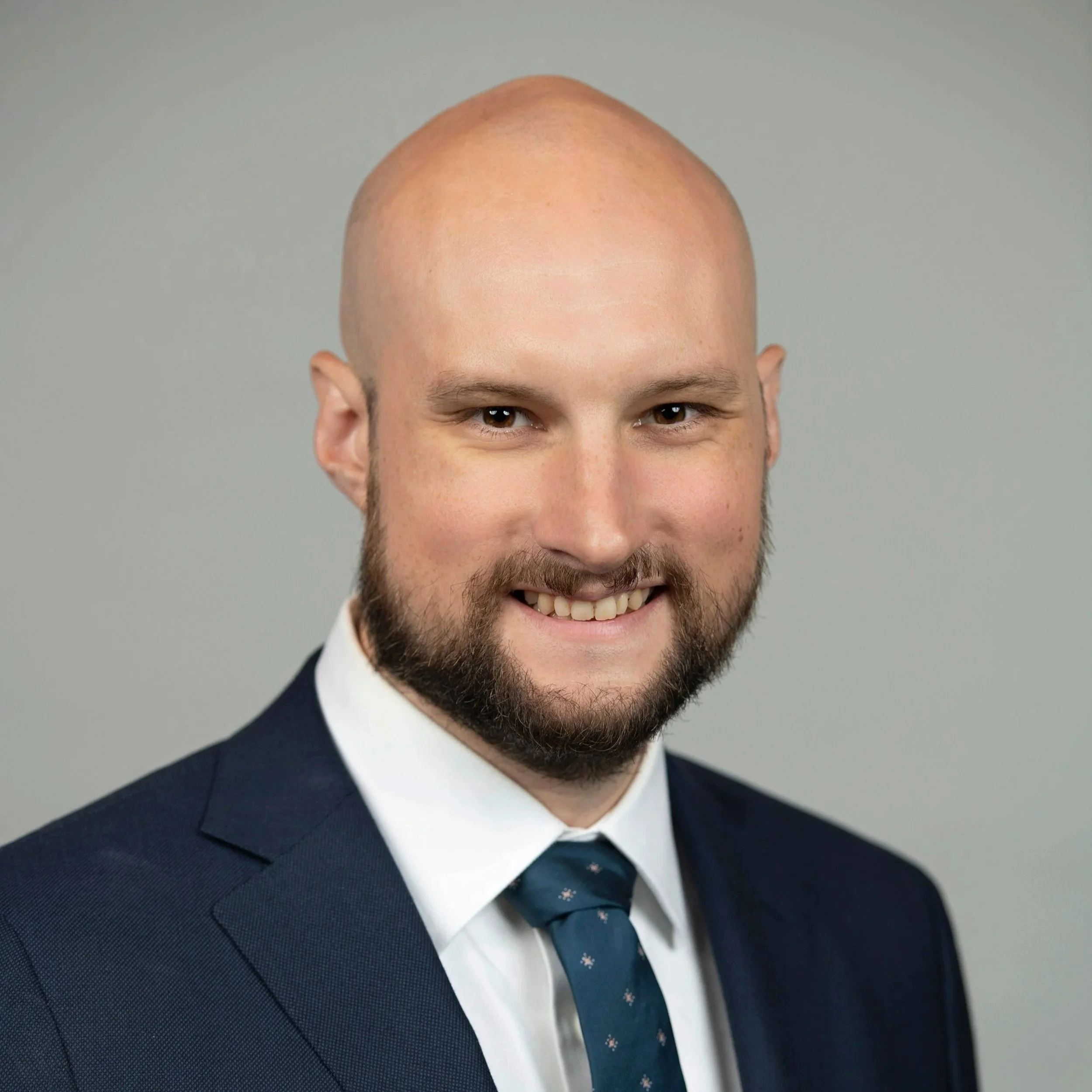 Close-up of a smiling man with a bald head, brown eyes, and facial hair, wearing a dark blue suit, white shirt, and a patterned blue tie, against a plain gray background.