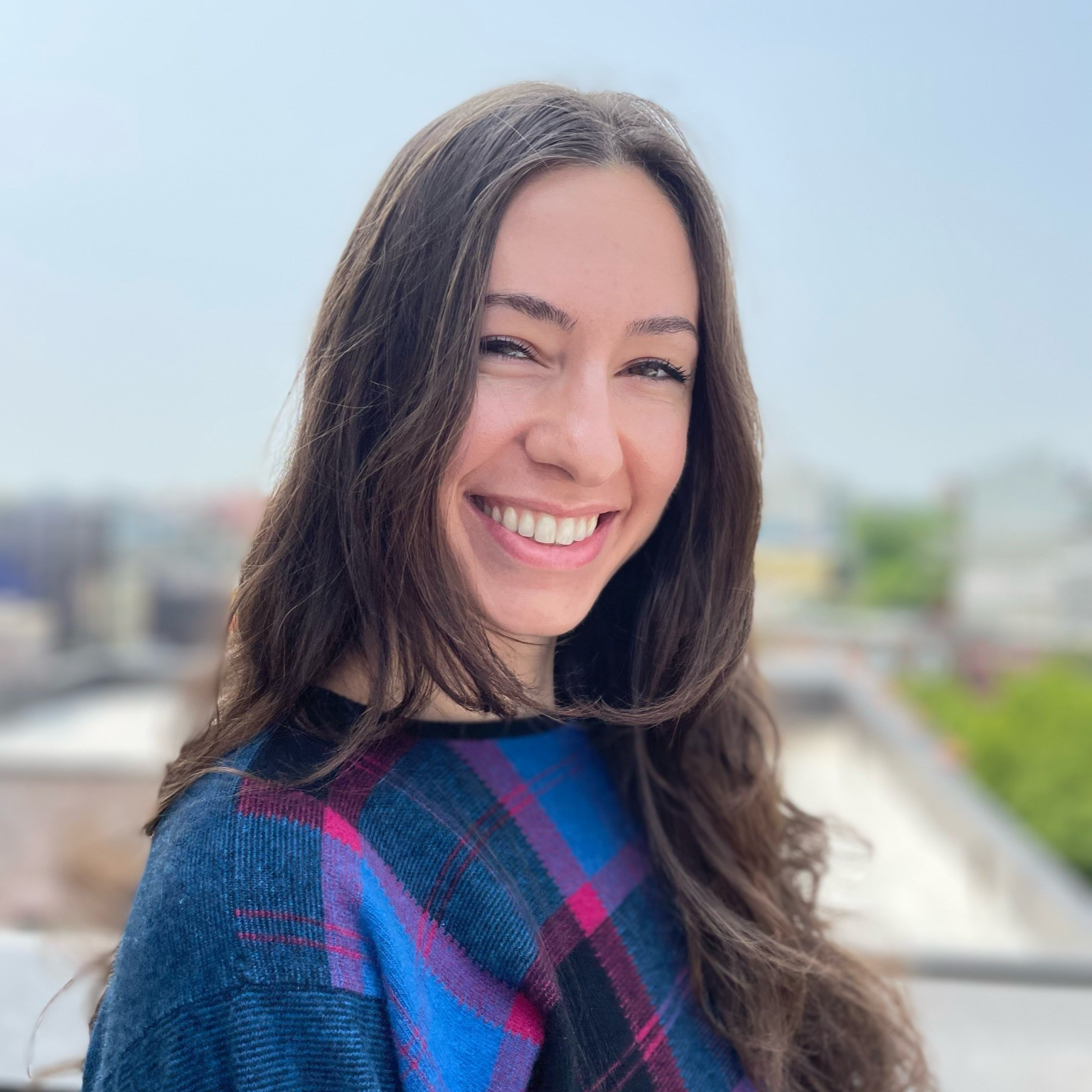 Smiling woman with long brown hair, wearing a blue and purple plaid shirt, standing outdoors with an urban background.