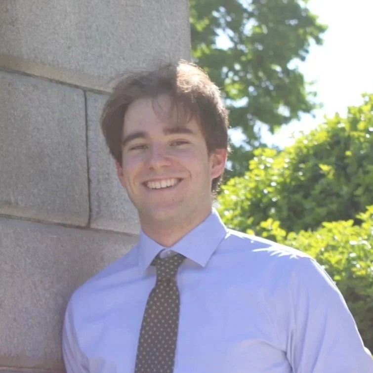 Young man with dark hair, smiling, wearing a light blue dress shirt and a dark polka dot tie, standing outdoors next to a concrete wall with green trees in the background.