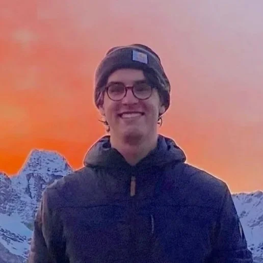 A young man with glasses and a beanie hat standing outdoors in front of snow-capped mountains during a colorful sunset.