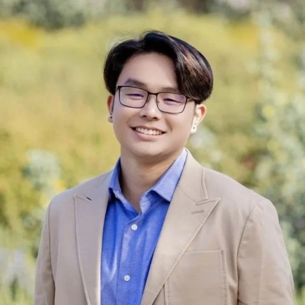 Young man with glasses, wearing a tan blazer over a blue shirt, smiling outdoors with blurred greenery in the background.