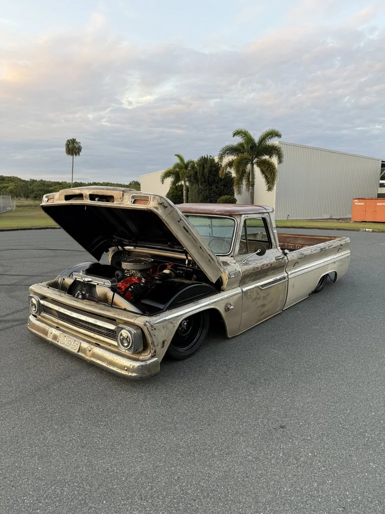 A vintage, rusty pickup truck with a weathered metal exterior, open hood revealing the engine, and lowered stance parked on a paved surface outdoors with palm trees and industrial buildings in the background under a cloudy sky.