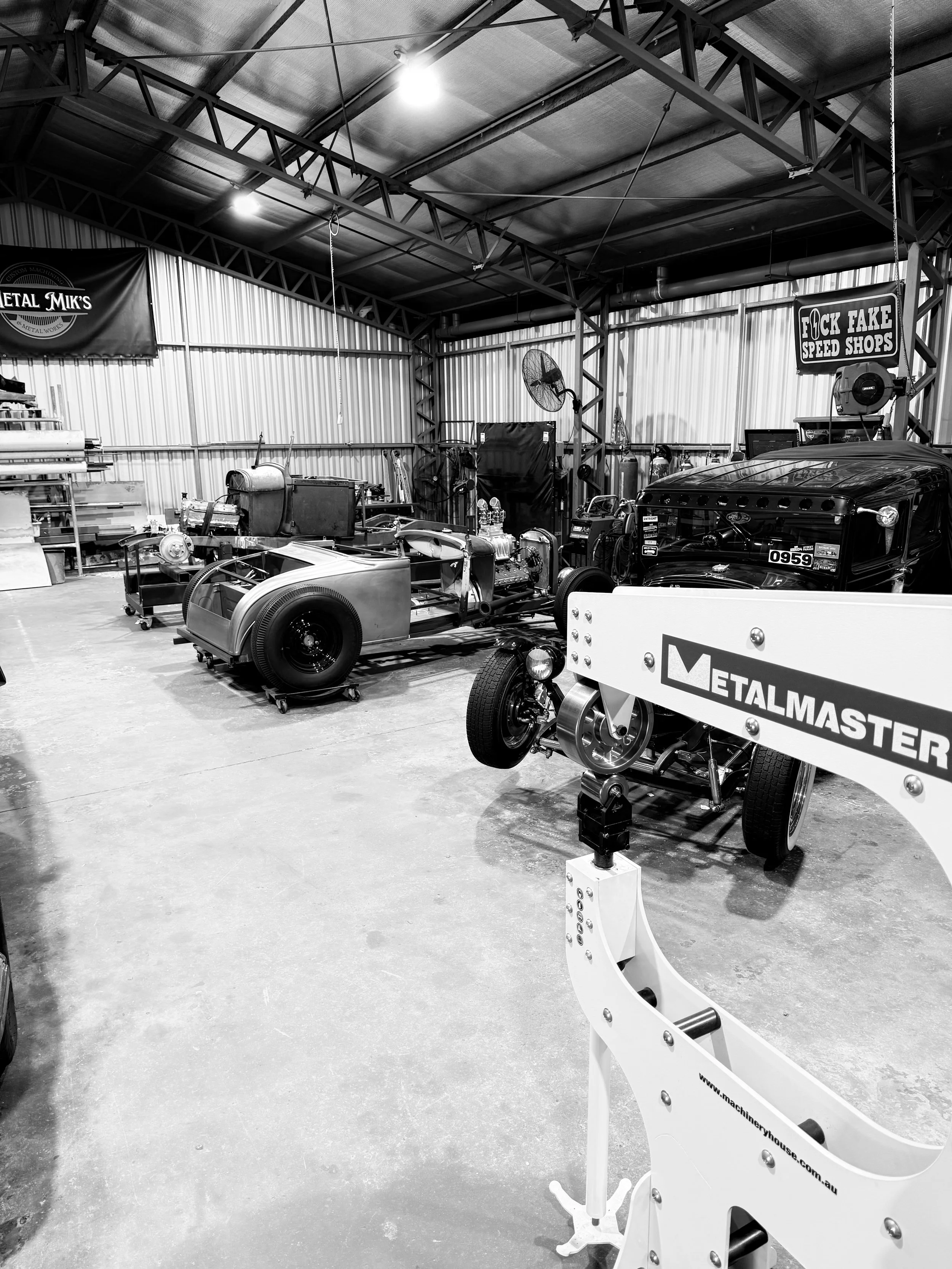 Black and white photo of a garage with restoration cars, tools, and equipment, including a partially assembled vintage vehicle, a wide fan, and signage with brand names like 'MetalMaster' and 'Metal Mik's', and a sign that reads 'F*** Fake Speed Shops'.