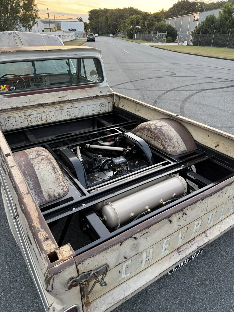 The back of a vintage pickup truck with its bed removed, showing a racing engine and a large silver air tank inside the truck bed area.