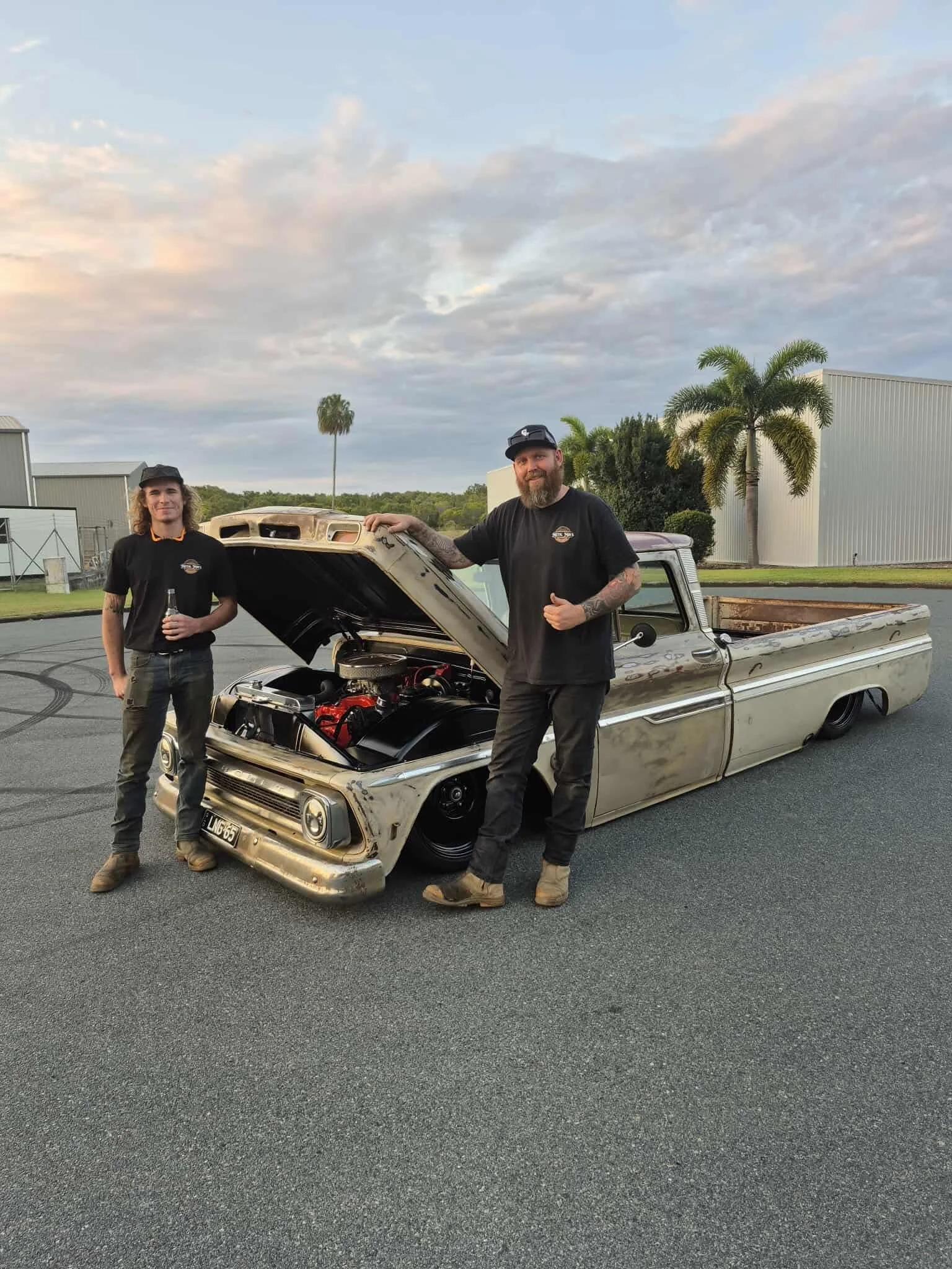 Two men standing in front of a vintage pickup truck with an open hood, possibly after working on or inspecting it, outdoors during sunset or late afternoon. Both men are dressed in black shirts and boots, with one holding a drink.