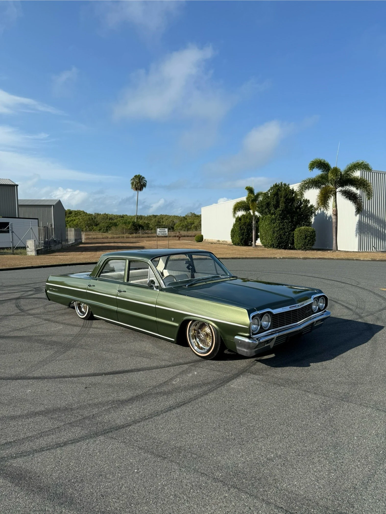 A vintage green and black classic car parked on an asphalt parking lot with tire marks. The background features palm trees, a blue sky with some clouds, and industrial buildings.