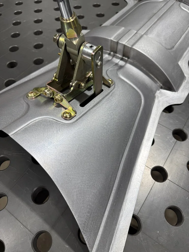 A close-up view of an aircraft control surface hinge mechanism attached to a metallic fuselage part, with a perforated metal work table underneath.