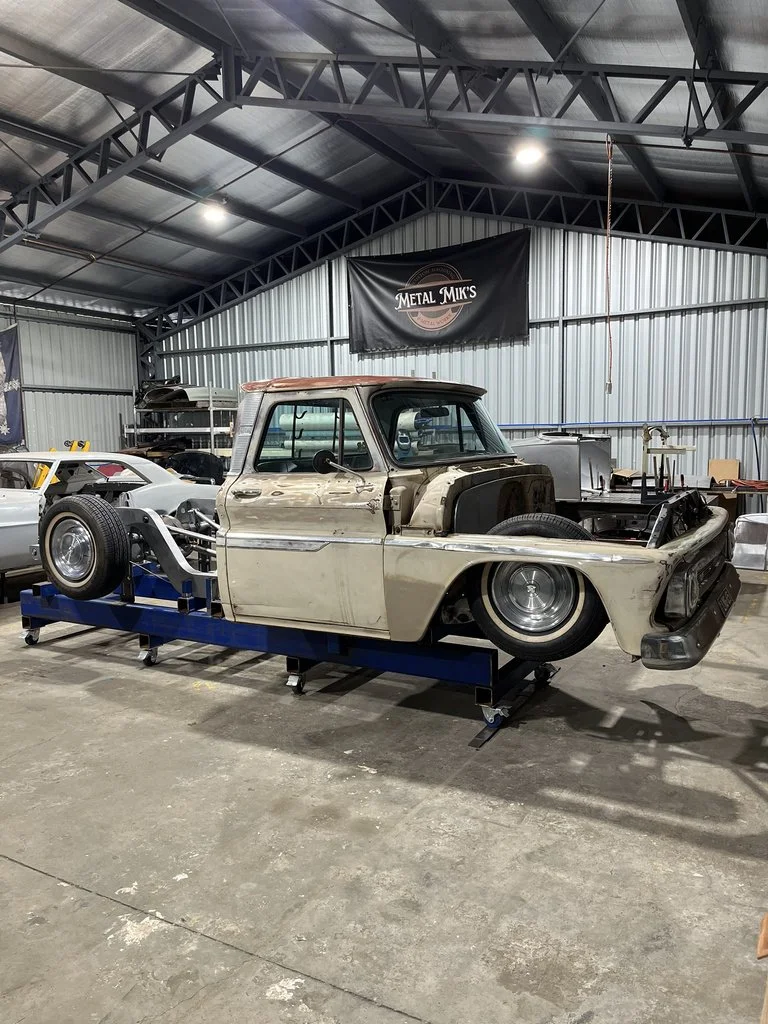 A vintage car body being restored inside a workshop, with car parts and tools around. The workspace has a metal roof and walls, with a banner that reads 'Metal MIKS' hanging on the back wall.