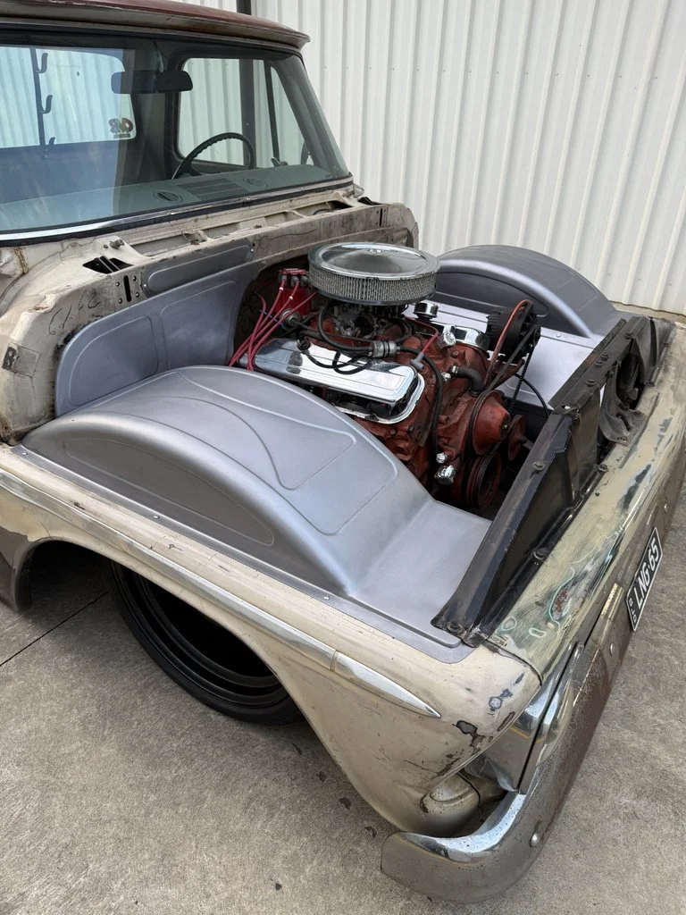 A vintage car with an exposed engine in the front, showing a red engine block, chrome air filter, and valve covers, with a damaged and weathered body.
