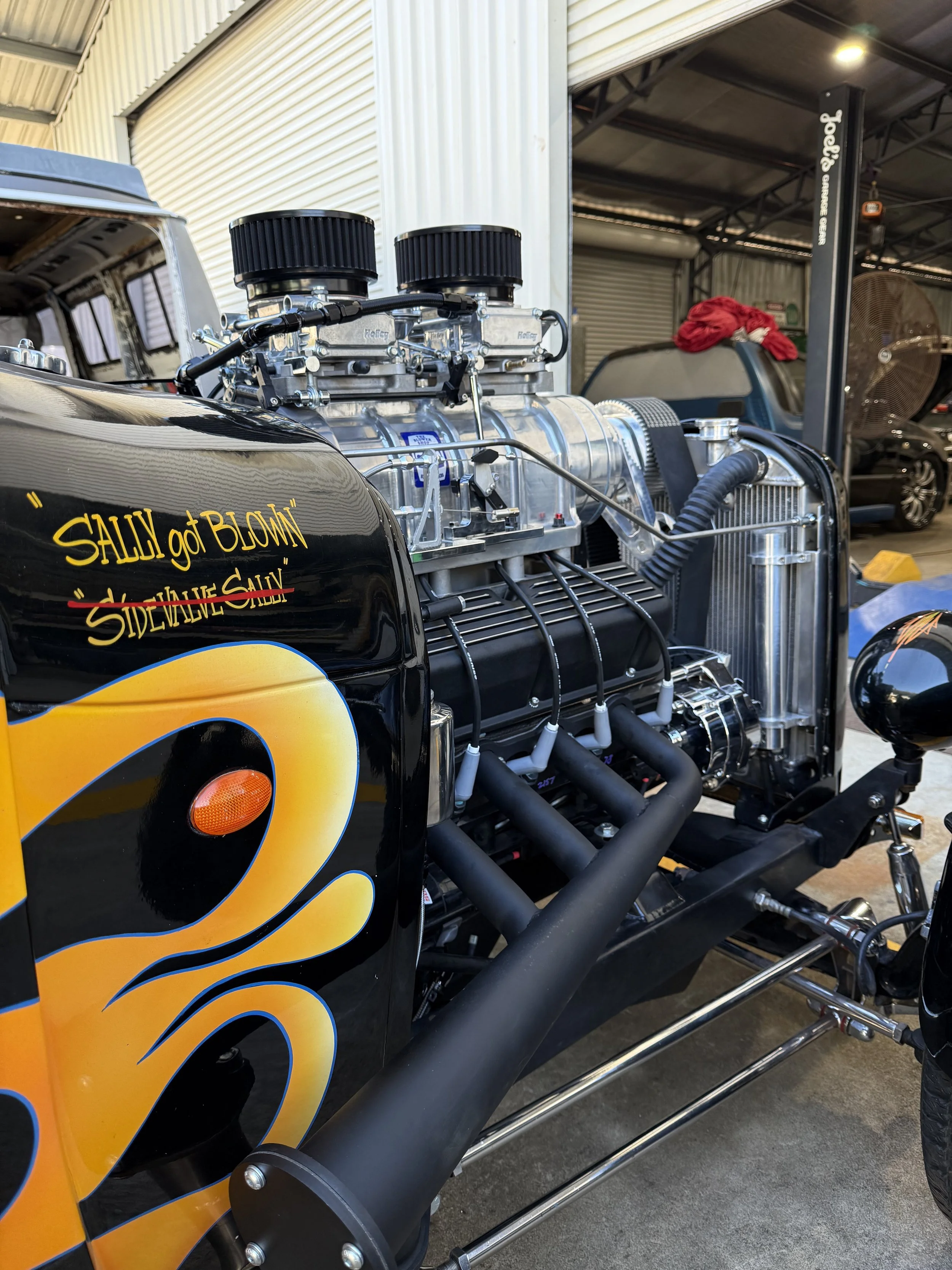 Close-up of a vintage race car engine with black exhaust pipes, positioned inside a garage with other vehicles and equipment in the background.