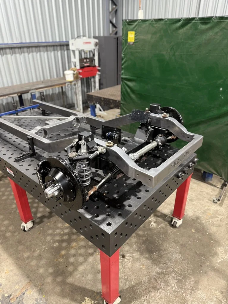 A metal engineering jig or fixture with springs, bolts, and rods, mounted on a black perforated work table with red legs in an industrial workshop.