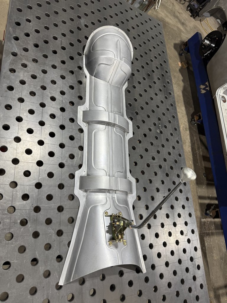 A metallic, machined aluminum component with a handle situated on a perforated metal work table.